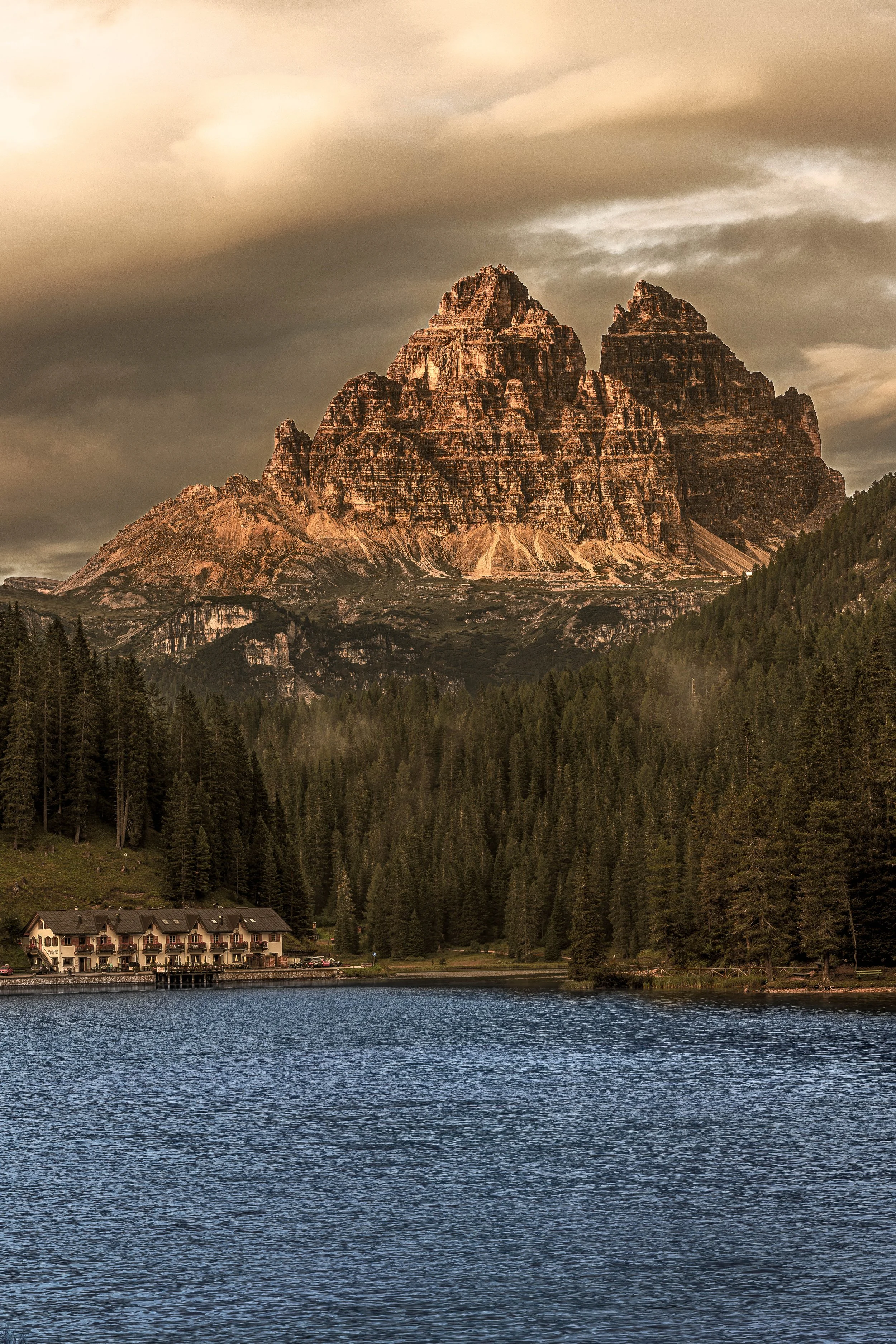 A large mountain with steep cliffs behind a forested area near a body of water, with a cluster of buildings at the water's edge and dark, cloudy sky.