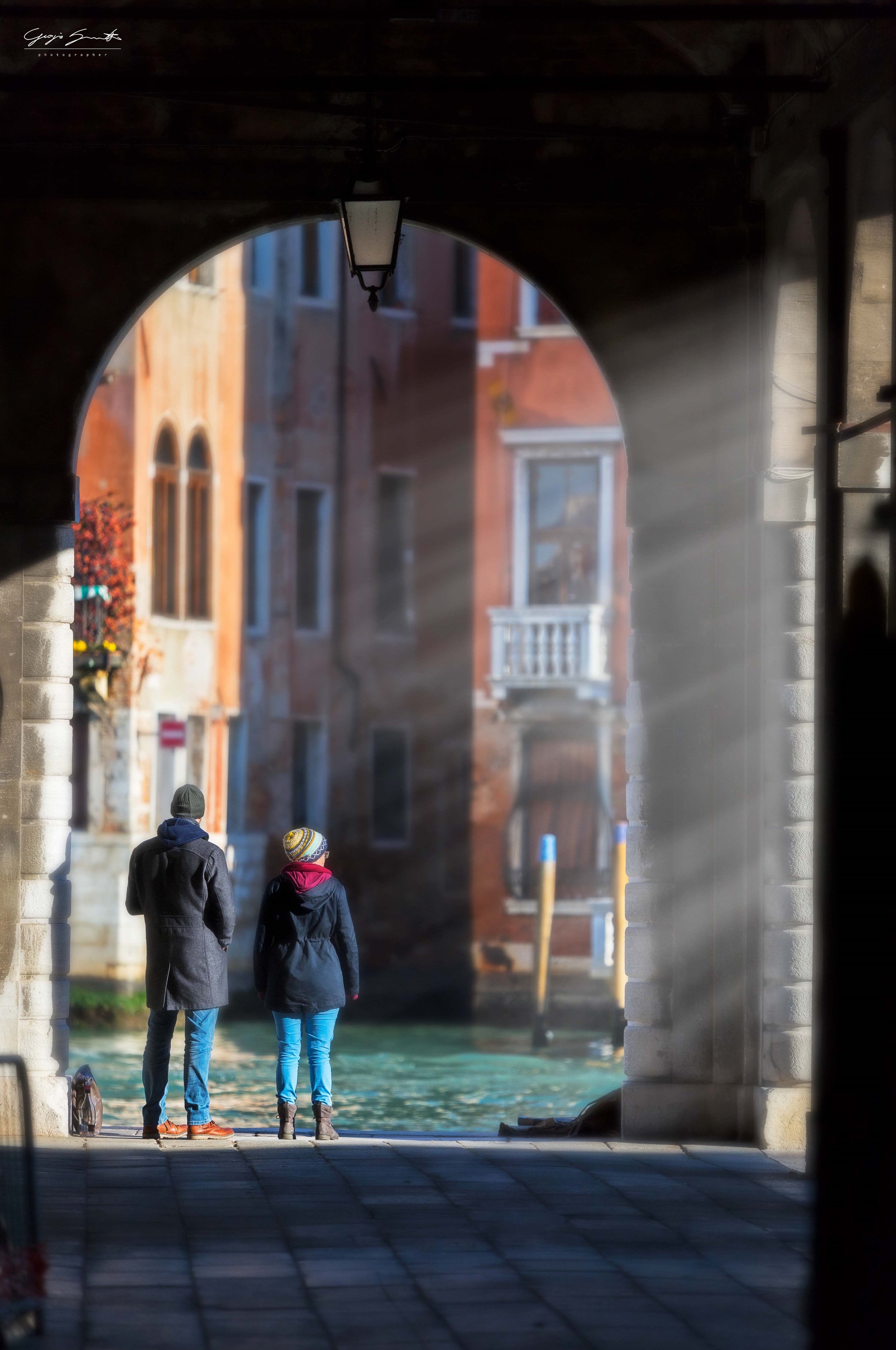 Two people walking along a canal in Venice, Italy, as viewed through an arched stone bridge.