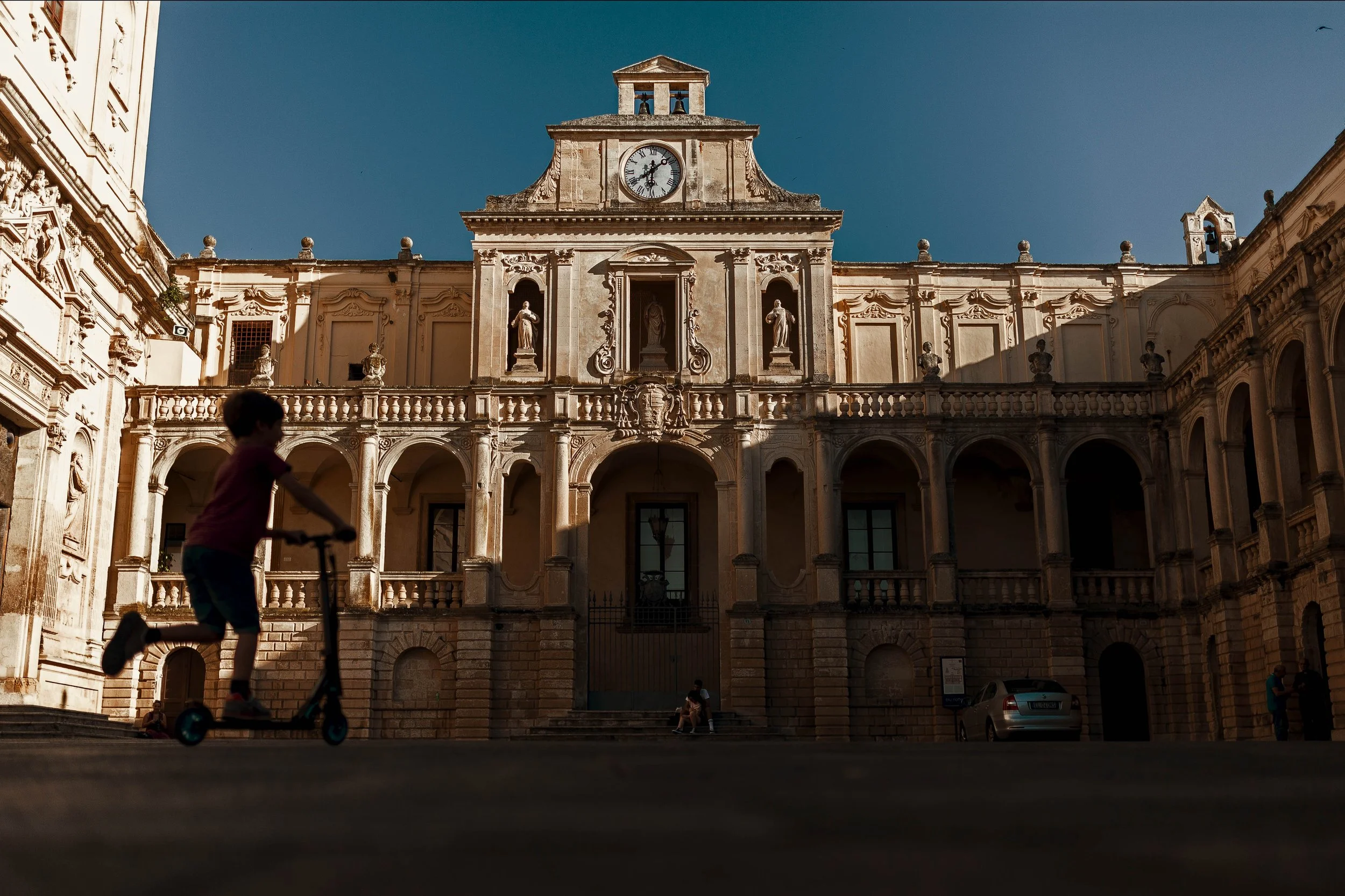 Street photography in a sunlit southern Italian Baroque piazza — a child on a scooter blurs past an ornate cathedral facade bathed in warm afternoon light under a deep blue sky. Candid urban moment, contrast of movement and timeless stone.