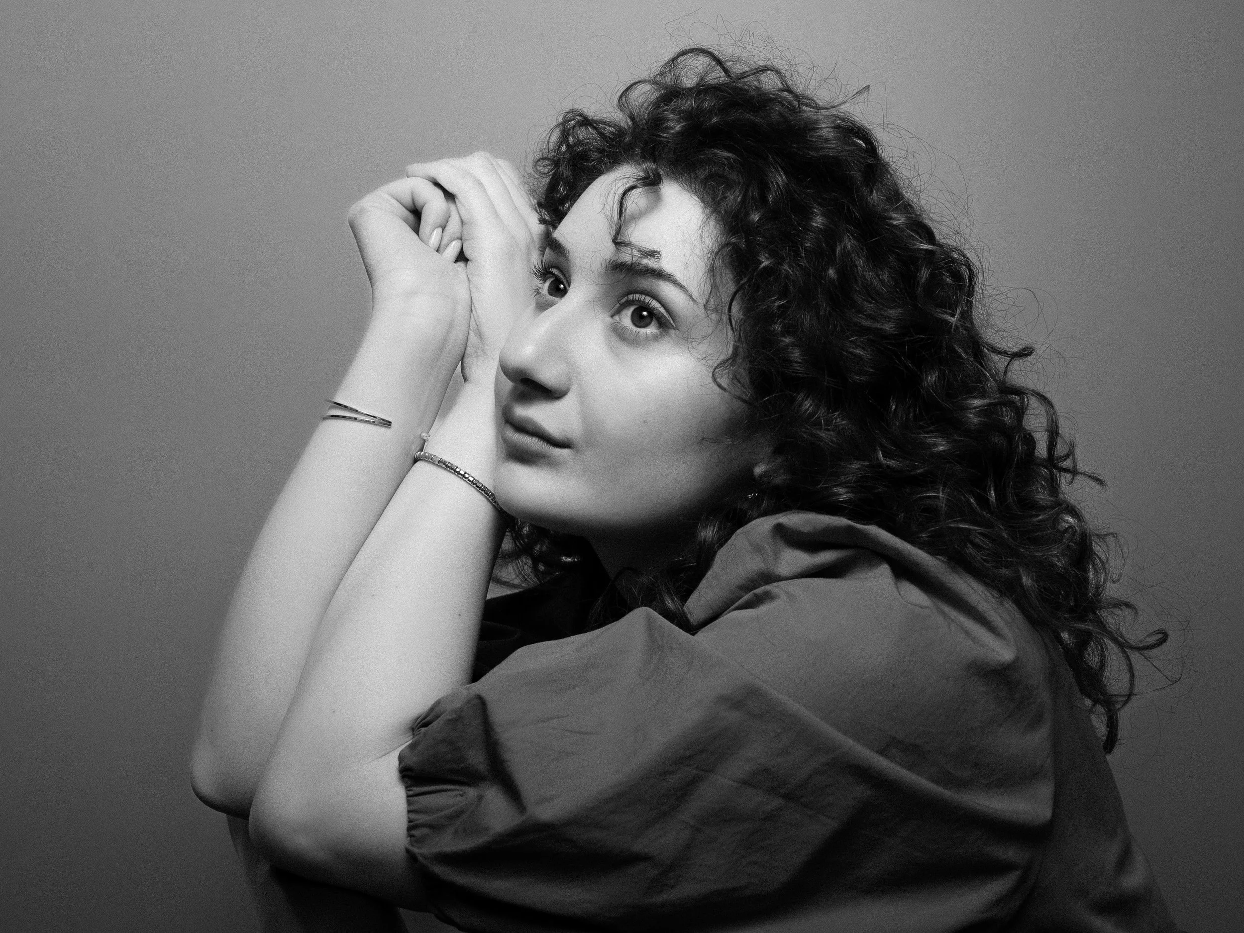 A young woman with curly hair resting her arms on her head, looking up, in black and white, with a plain background.