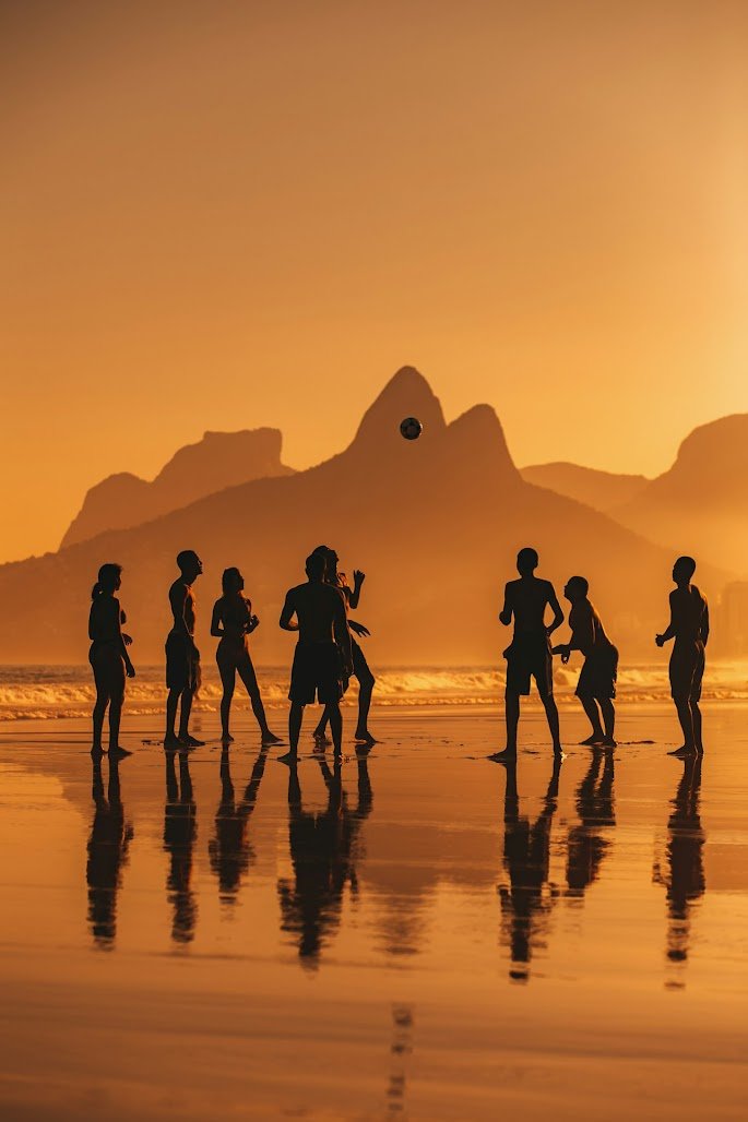 Silhouettes of children playing with a ball on the beach at sunset, with mountains in the background and their reflections on the wet sand.