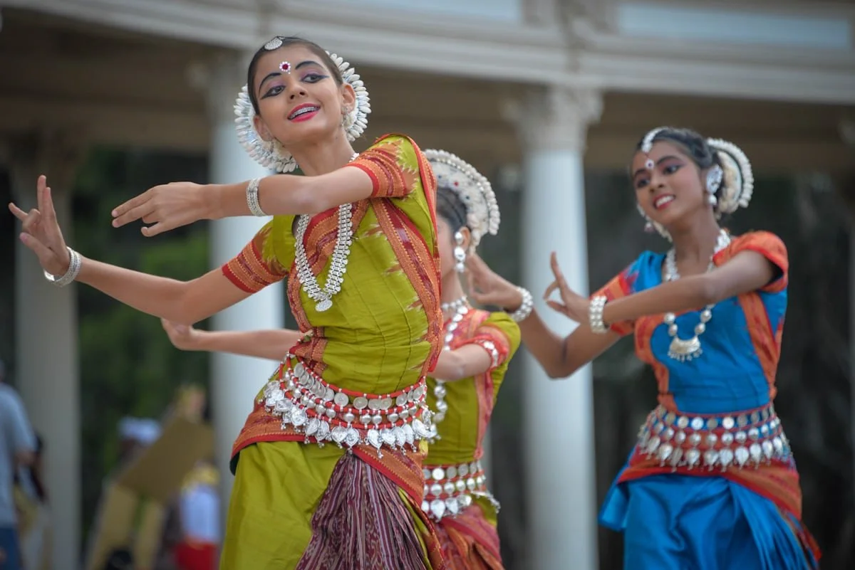 Young girls wearing traditional colorful Indian attire and jewelry, dancing outdoors with a pavilion and columns in the background.