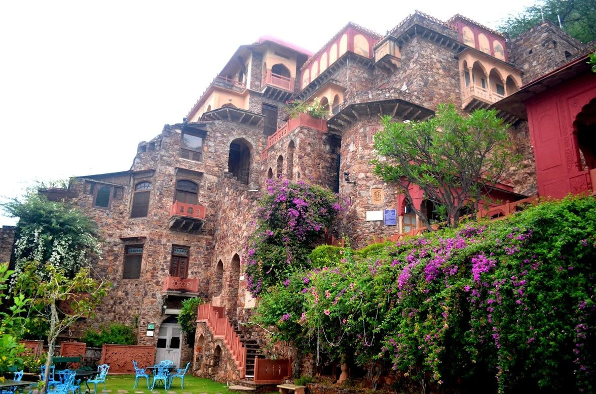 A multi-story stone castle-like building with arched windows and balconies, surrounded by lush green bushes and purple flowering plants, with outdoor seating and a staircase leading up to it.