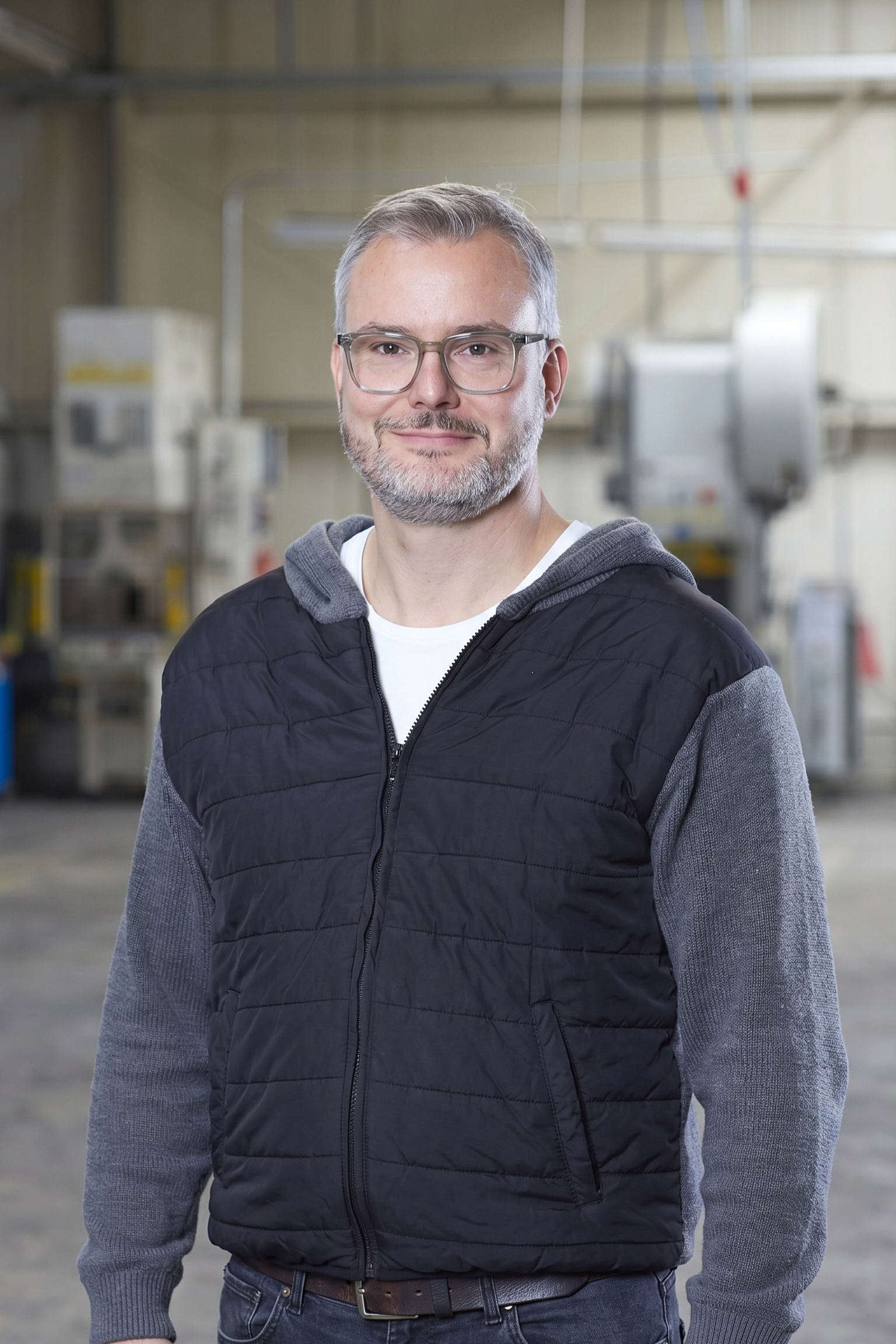 A man with glasses and gray hair standing in an industrial setting, wearing a black quilted jacket over a gray hoodie, smiling.