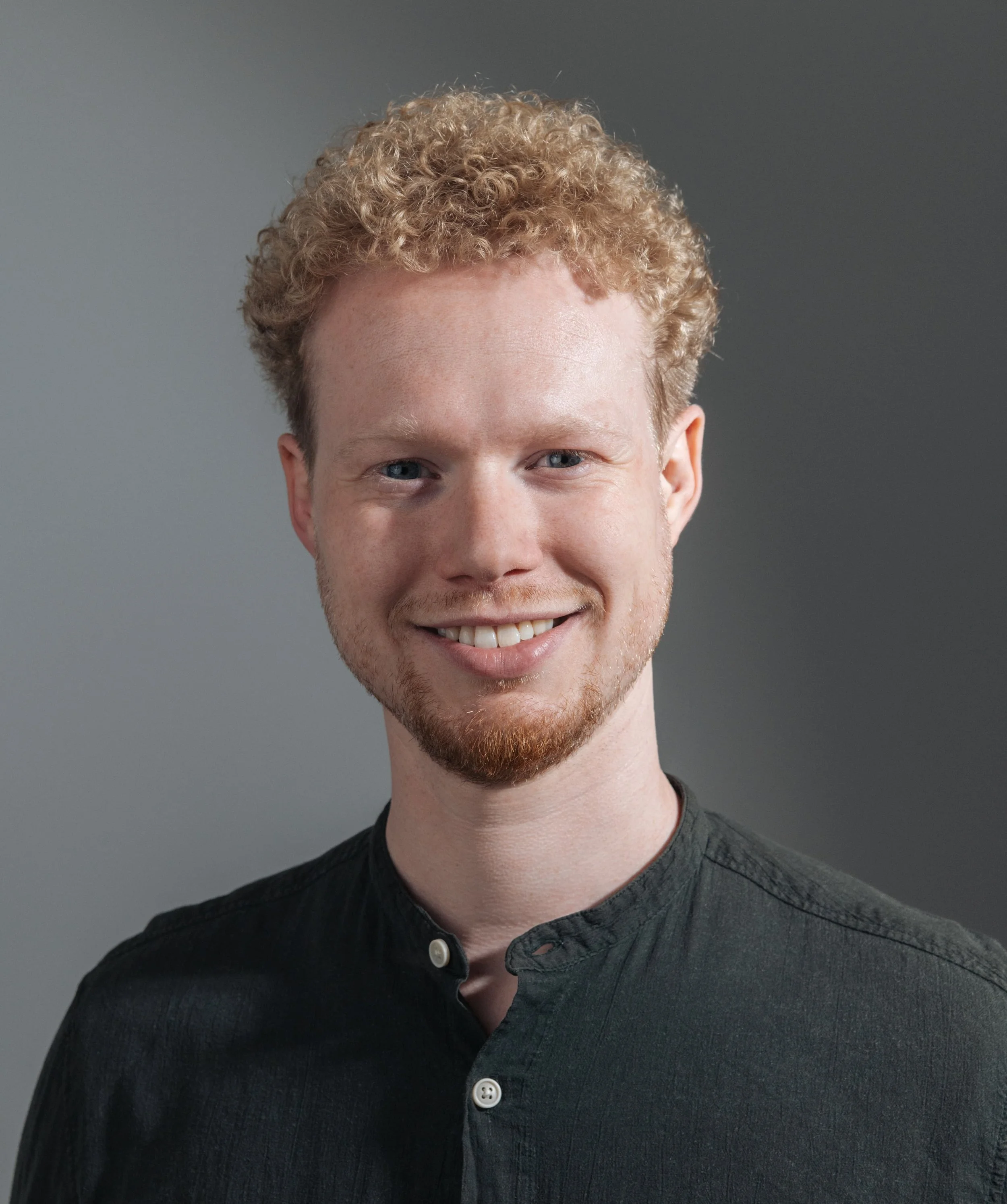 Close-up of a smiling young man with light skin, curly red hair, and blue eyes, wearing a black button-up shirt against a plain gray background.
