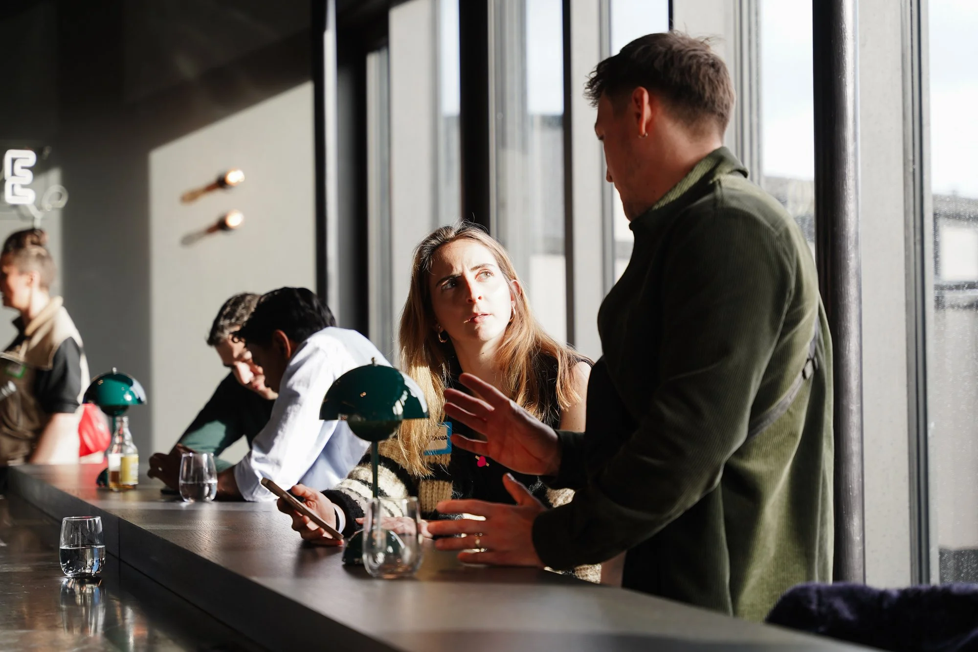 A woman with long hair and earrings sitting at a counter, looking up at a man standing next to her, who is gesturing with his hands. There are other people sitting at the counter, with glasses and lamps in front of them, inside a well-lit room with large windows.