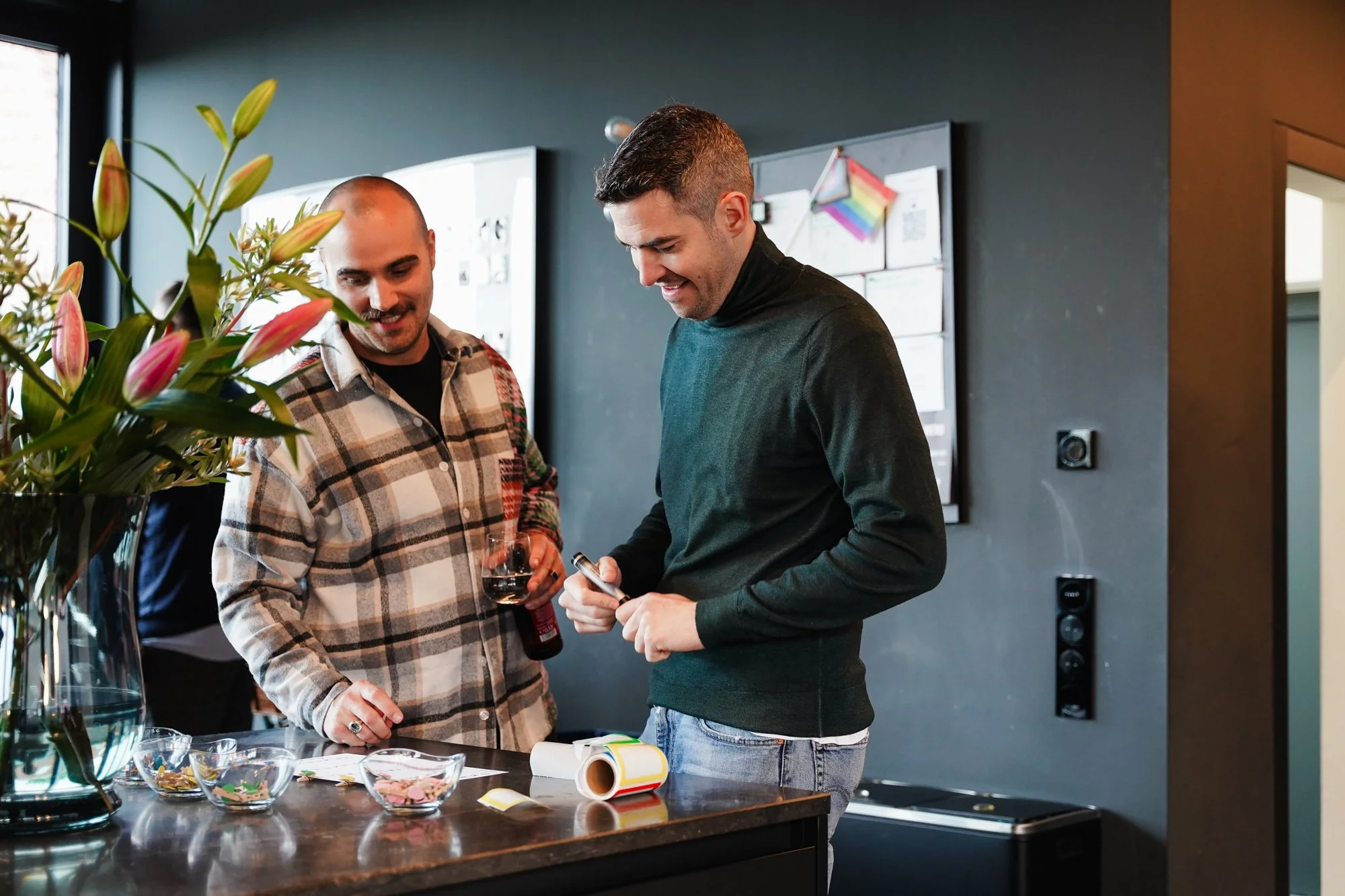 Two men are standing in a modern interior space, one is holding a glass of red wine and the other is writing on paper. There are various craft supplies and a roll of paper on the table in front of them, with a vase of pink lilies nearby. A chalkboard with colorful drawings and notes is on the wall behind them.