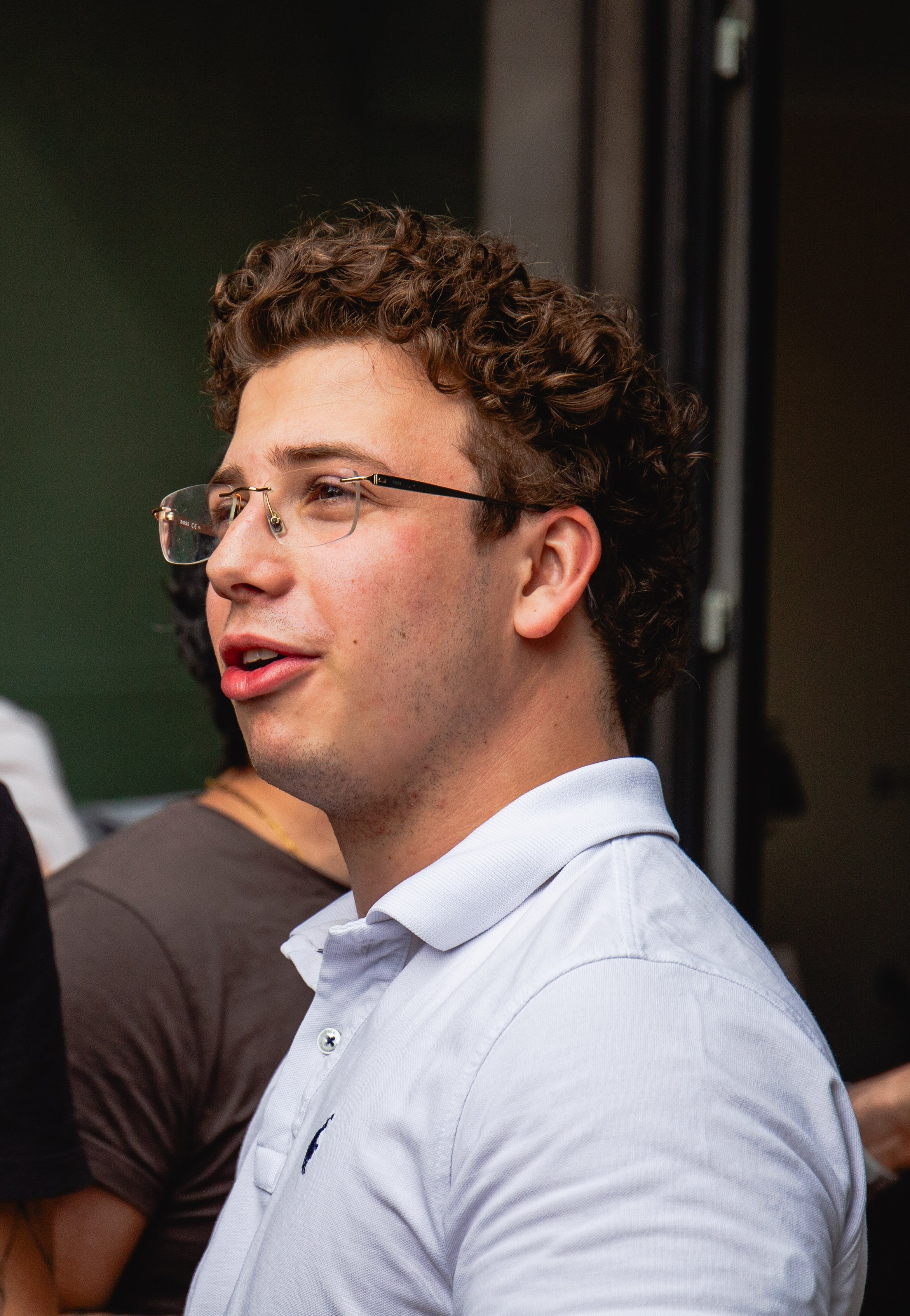 Close-up of a young man with curly brown hair, glasses, and a white collared shirt, smiling and looking to his right in a group setting.