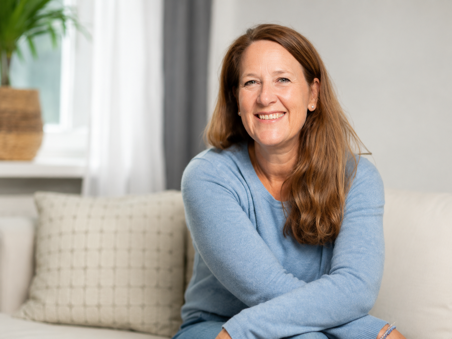 A woman with reddish-brown hair, smiling, wearing a light blue sweater, sitting on a beige couch with a pillow behind her, in a bright room with white curtains and a potted plant.