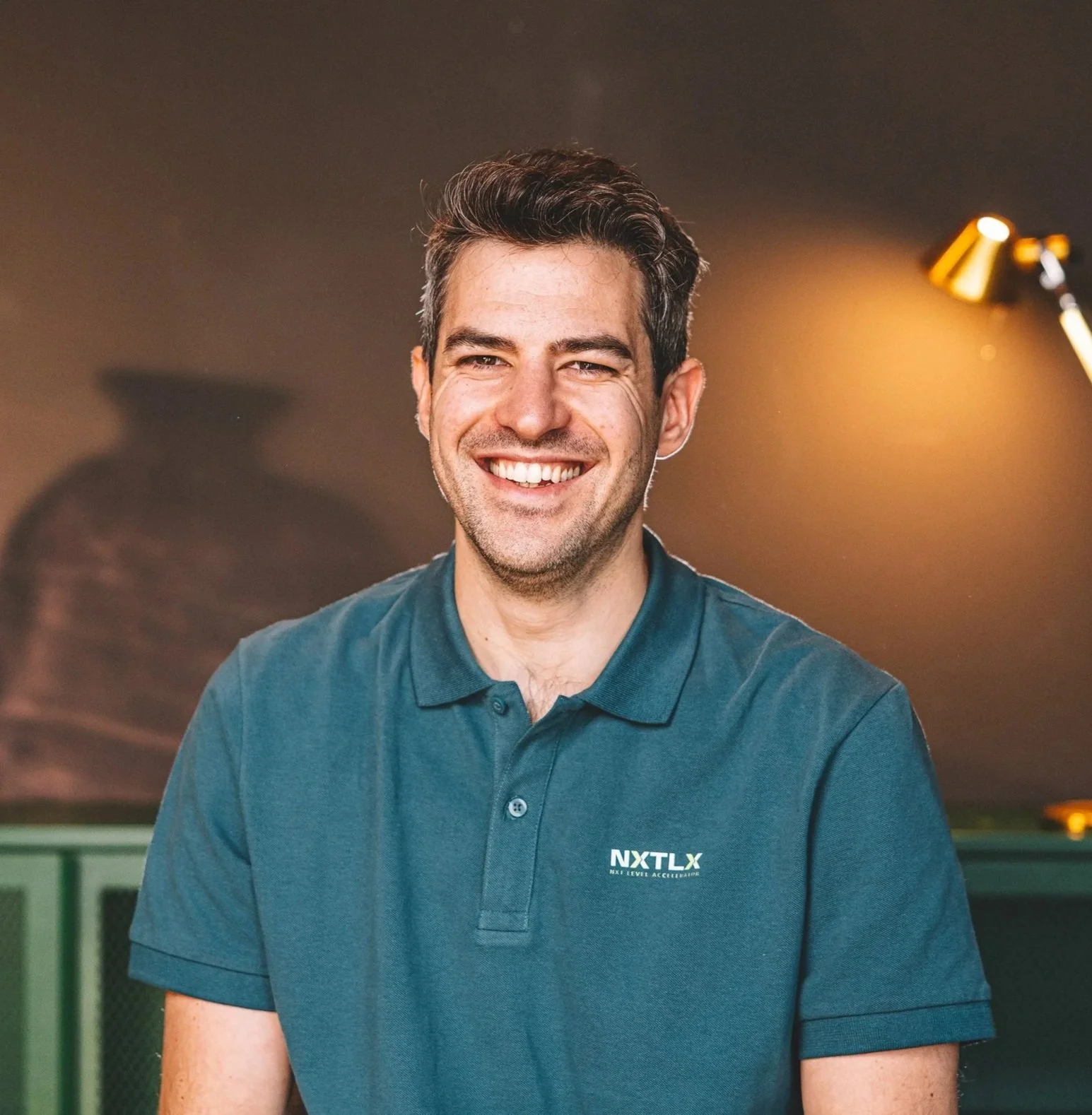 A smiling man with dark, wavy hair to the side, wearing a dark blue collared shirt with a logo that reads 'NXTLX' on the chest, sitting in front of a background with warm lighting and a blurred hint of other people.