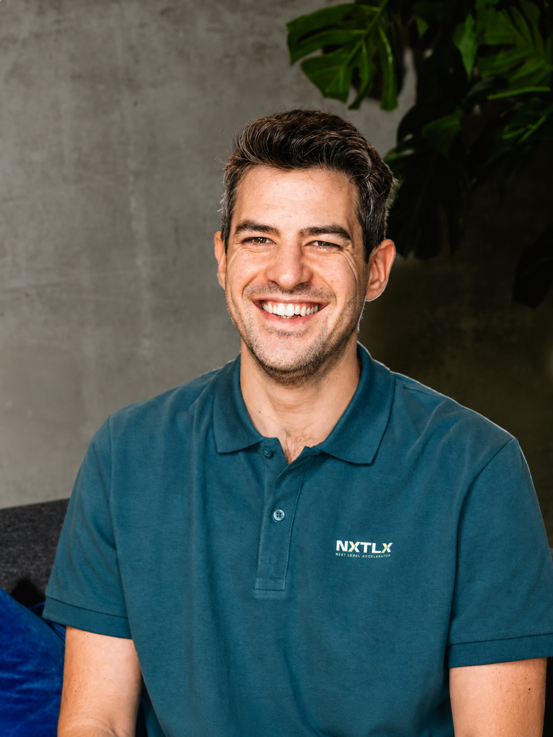 A smiling man with dark, wavy hair to the side, wearing a dark blue collared shirt with a logo that reads 'NXTLX' on the chest, sitting in front of a background with warm lighting and a blurred hint of other people.