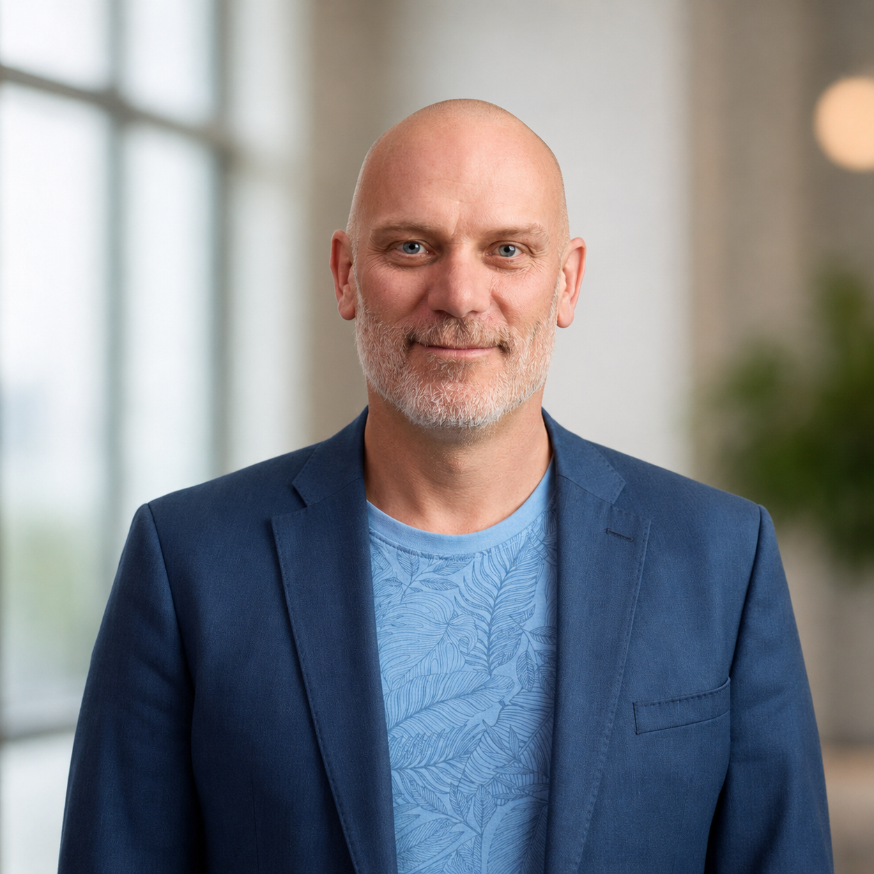A middle-aged man with a bald head and a gray beard, wearing a blue blazer and a light blue shirt with a leaf pattern, standing indoors with a blurred background of windows and a plant.