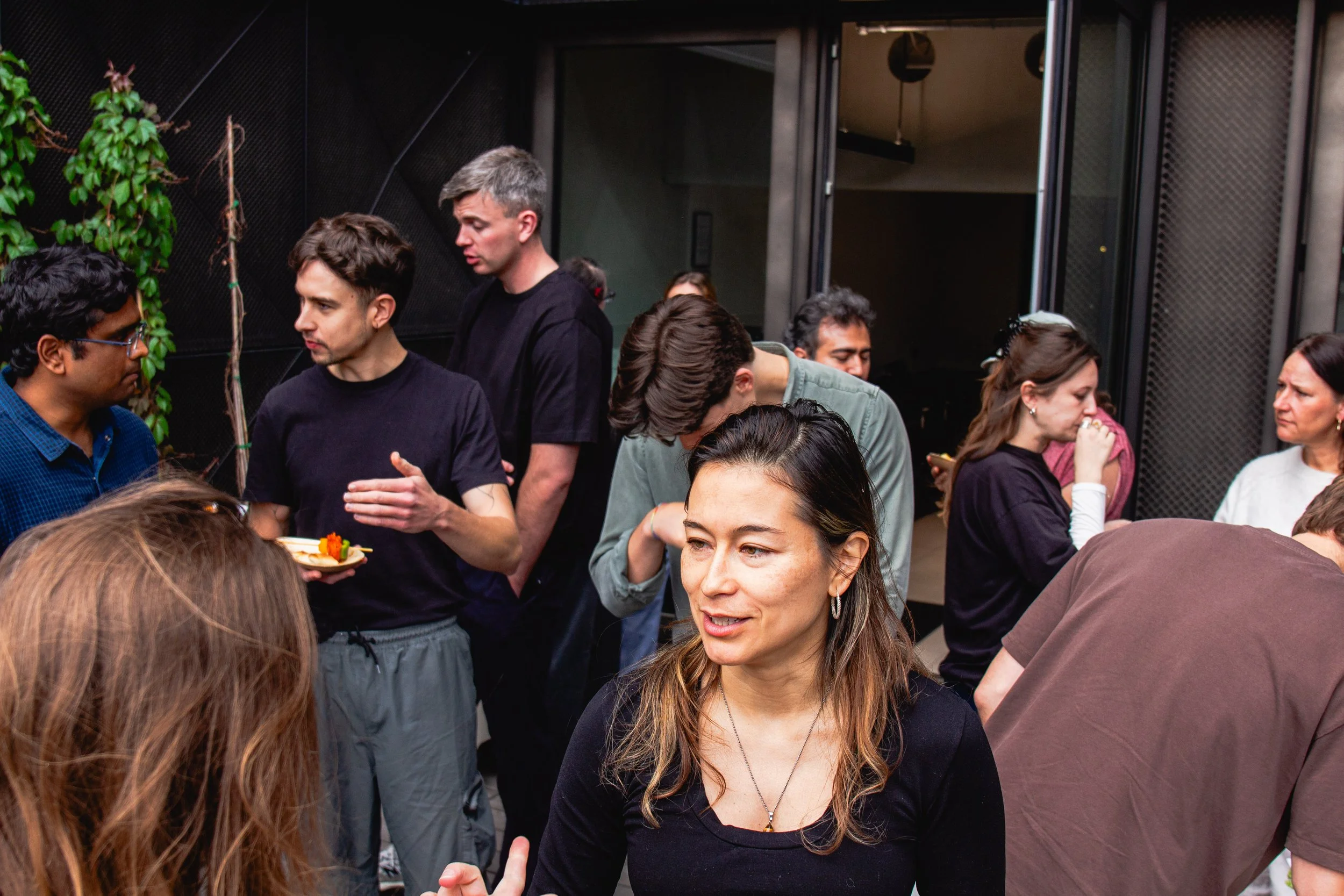 Group of people gathered indoors, some talking, others looking at their phones, with a plant and dark decor in the background.