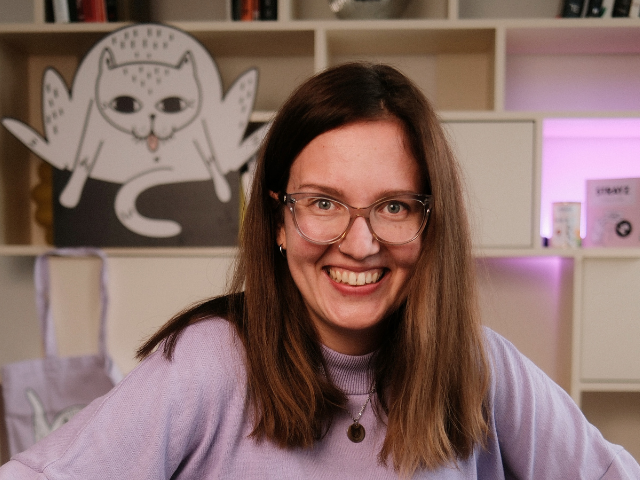 Woman with long brown hair and glasses smiling in a lavender turtleneck sweater, with a playful cat illustration and shelves in the background