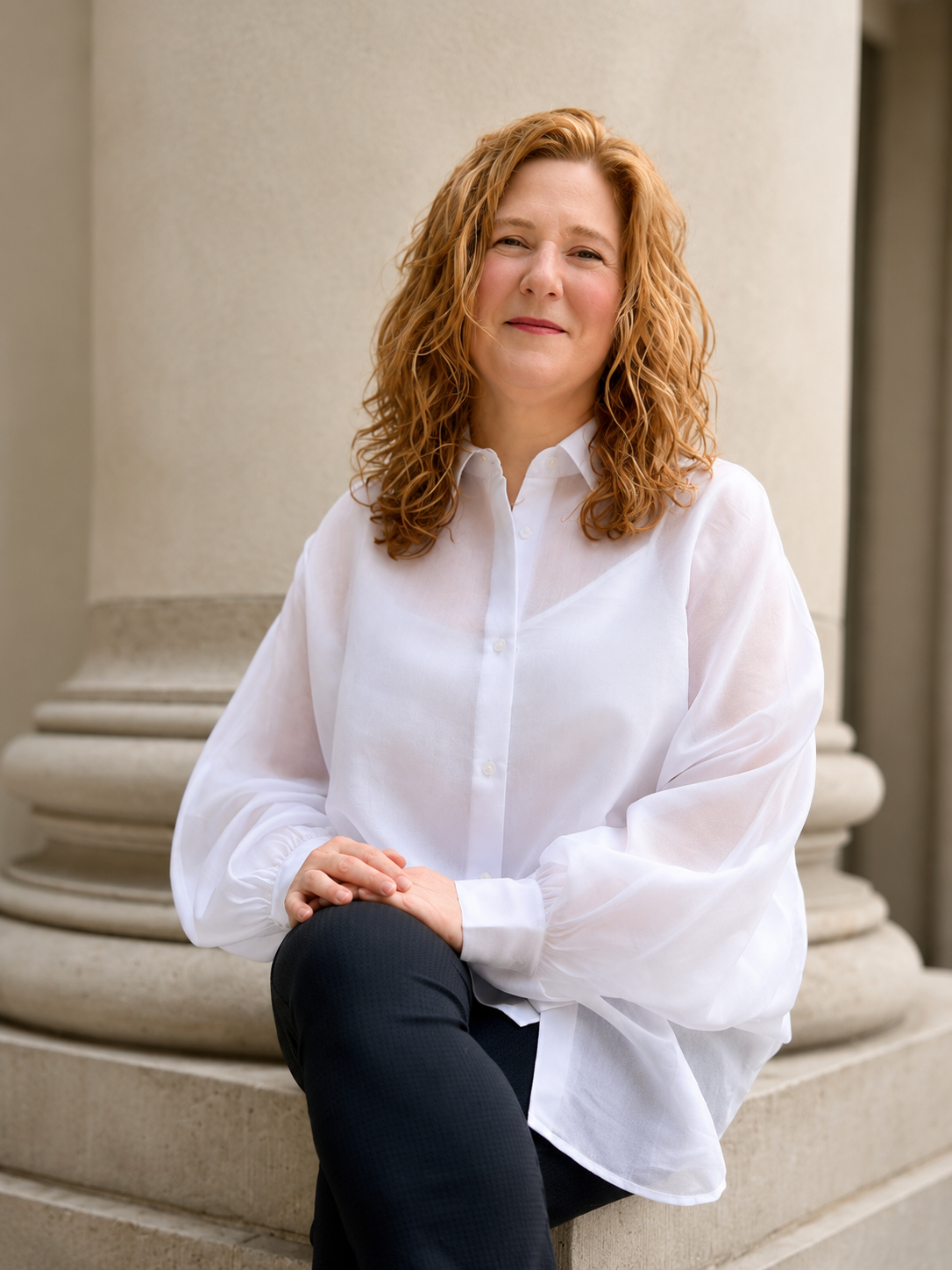 A woman with red, curly hair, wearing a white button-up shirt, sitting on the steps of a building with pillars.