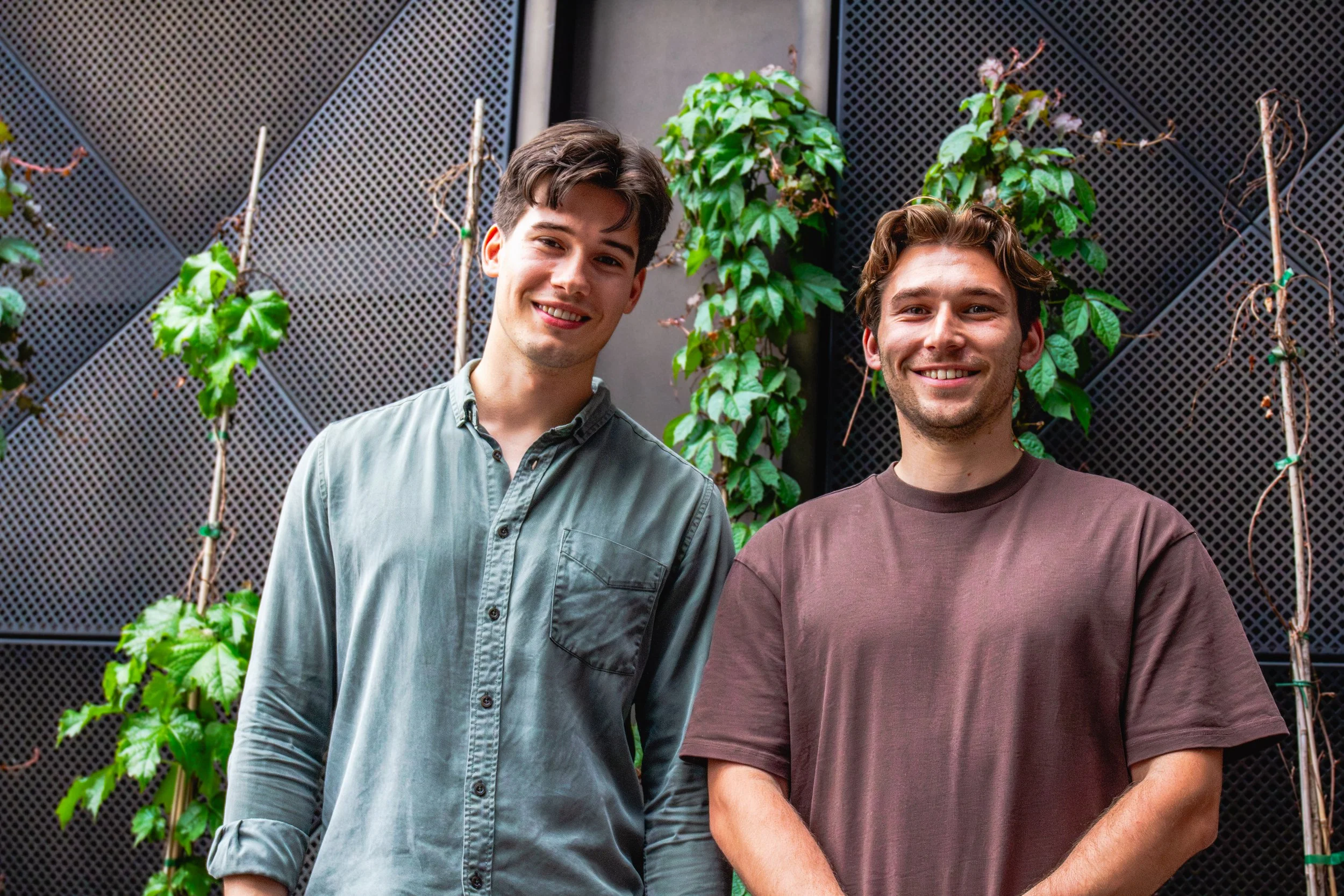 Two young men standing outdoors in front of a black mesh wall with green climbing plants. One is wearing a light green button-up shirt and the other a brown T-shirt. Both are smiling.