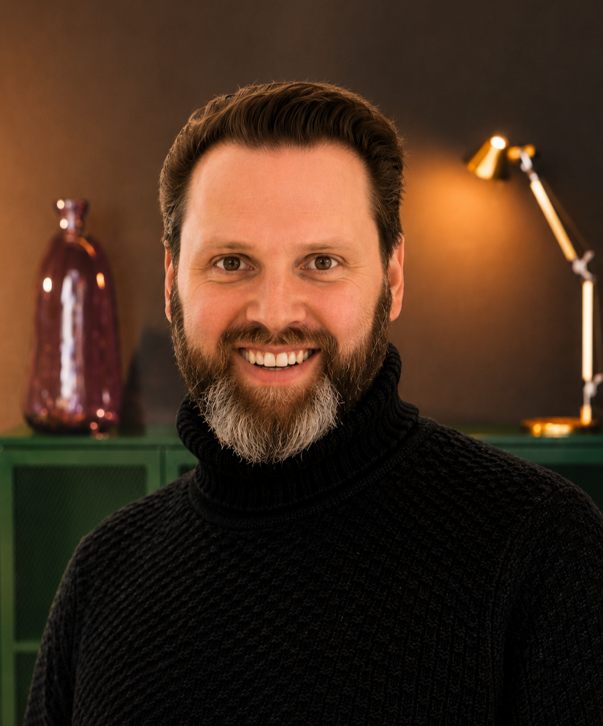 A smiling man with brown hair and a beard wearing a black turtleneck sweater, standing indoors with a green cabinet, a purple decorative vase, and a gold desk lamp in the background.