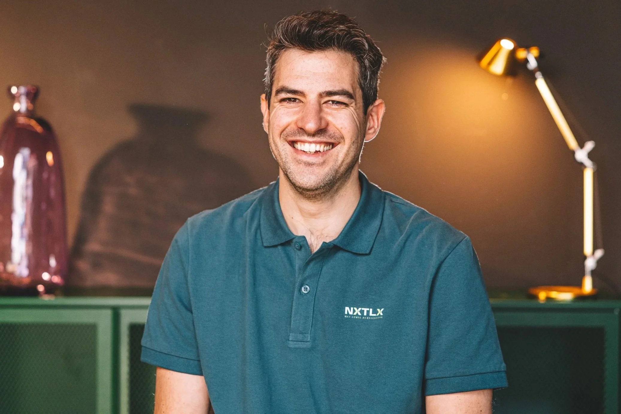 A smiling man with short dark hair wearing a teal polo shirt and sitting at a table in front of a ping pong table in an indoor gaming area.