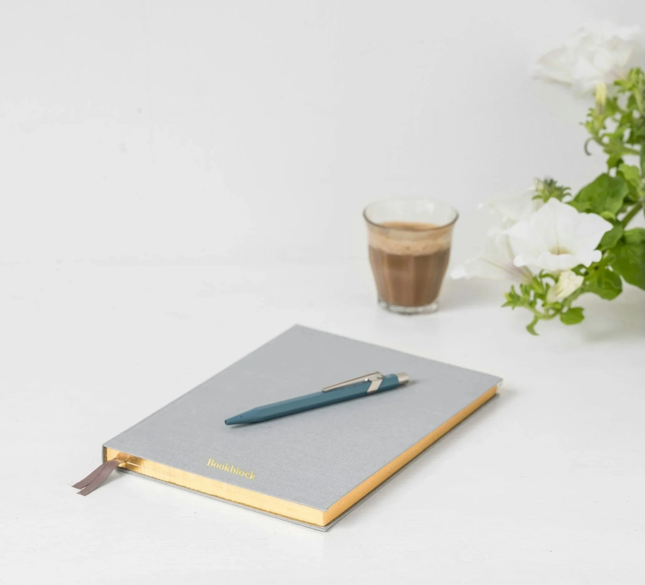 A gray book with a brown spine and a brown ribbon bookmark, lying on a gray surface. A blue pen and a yellow pencil are on top of the book. In the background, there are white flowers and green leaves, a clear glass cup with a brown drink, and a small gray notebook.