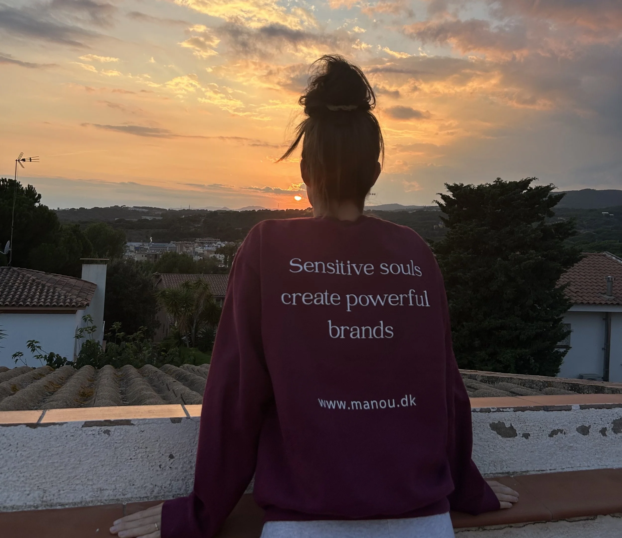Person with hair tied in a bun, wearing a maroon shirt that has white text, watching a sunset.