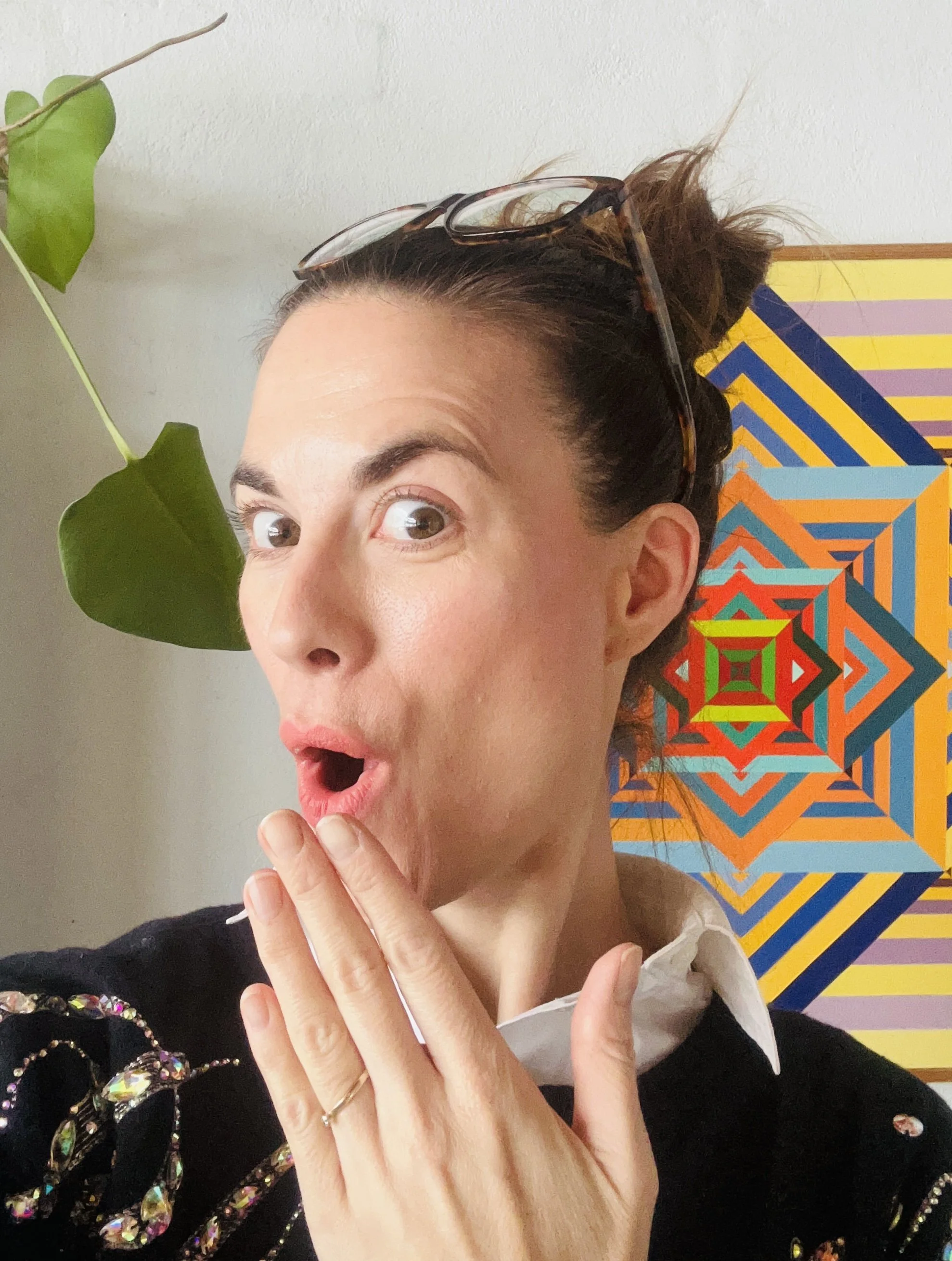 Surprising woman with glasses on her head, touched her face with fingers, against an abstract geometric colorful art background and a green plant.