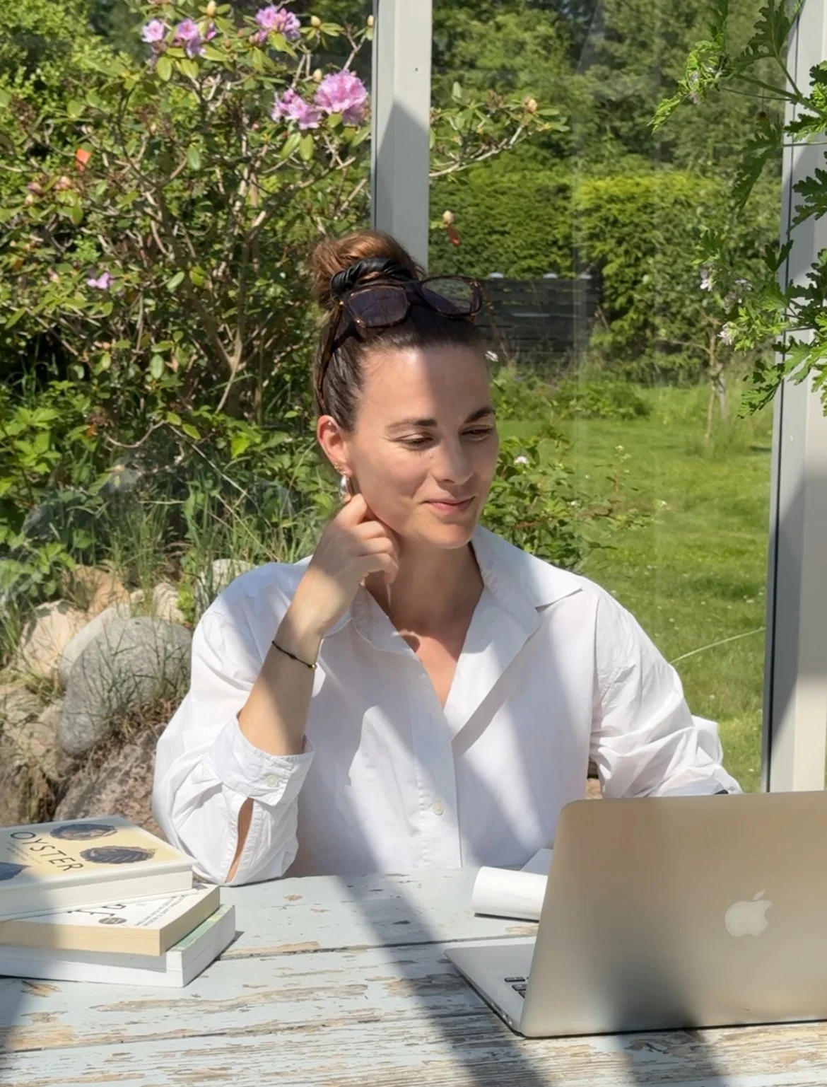 A young woman sitting at a white wooden table outdoors, working on a laptop. She has her hair in a bun with sunglasses on top of her head, and is wearing a white shirt. There are books on the table, and a lush garden with pink flowers and green foliage in the background.