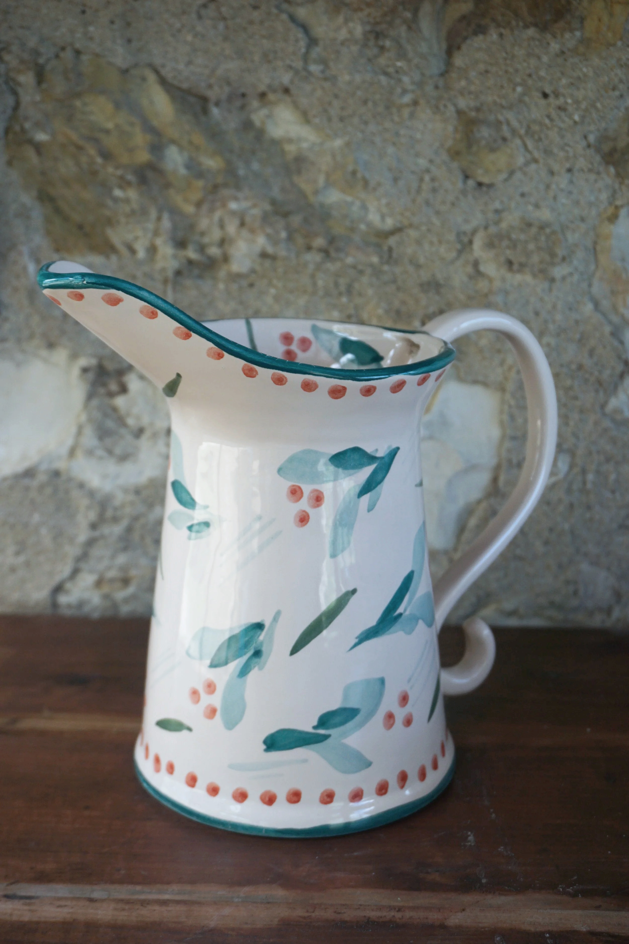 A ceramic pitcher with a floral design in shades of green and red, placed on a wooden surface with a stone wall in the background.