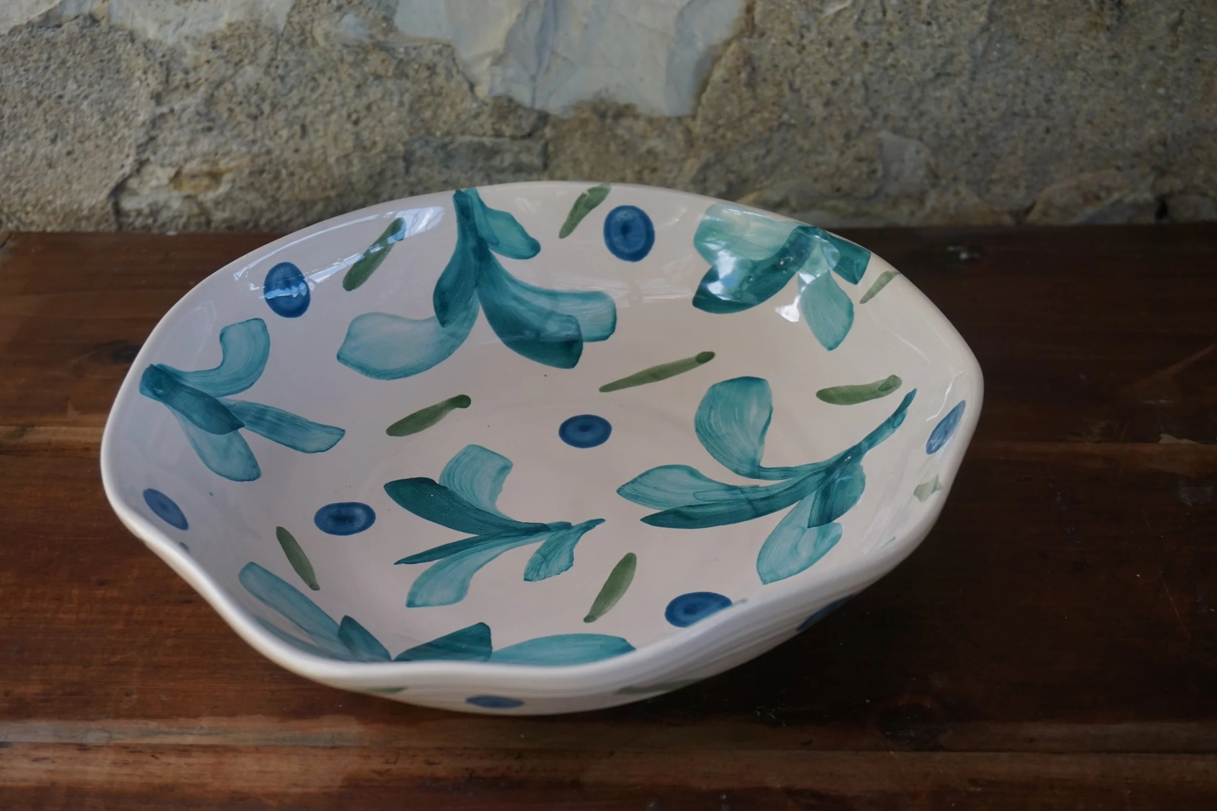 A white ceramic bowl decorated with blue and green brushstroke patterns, placed on a wooden surface against a stone wall background.