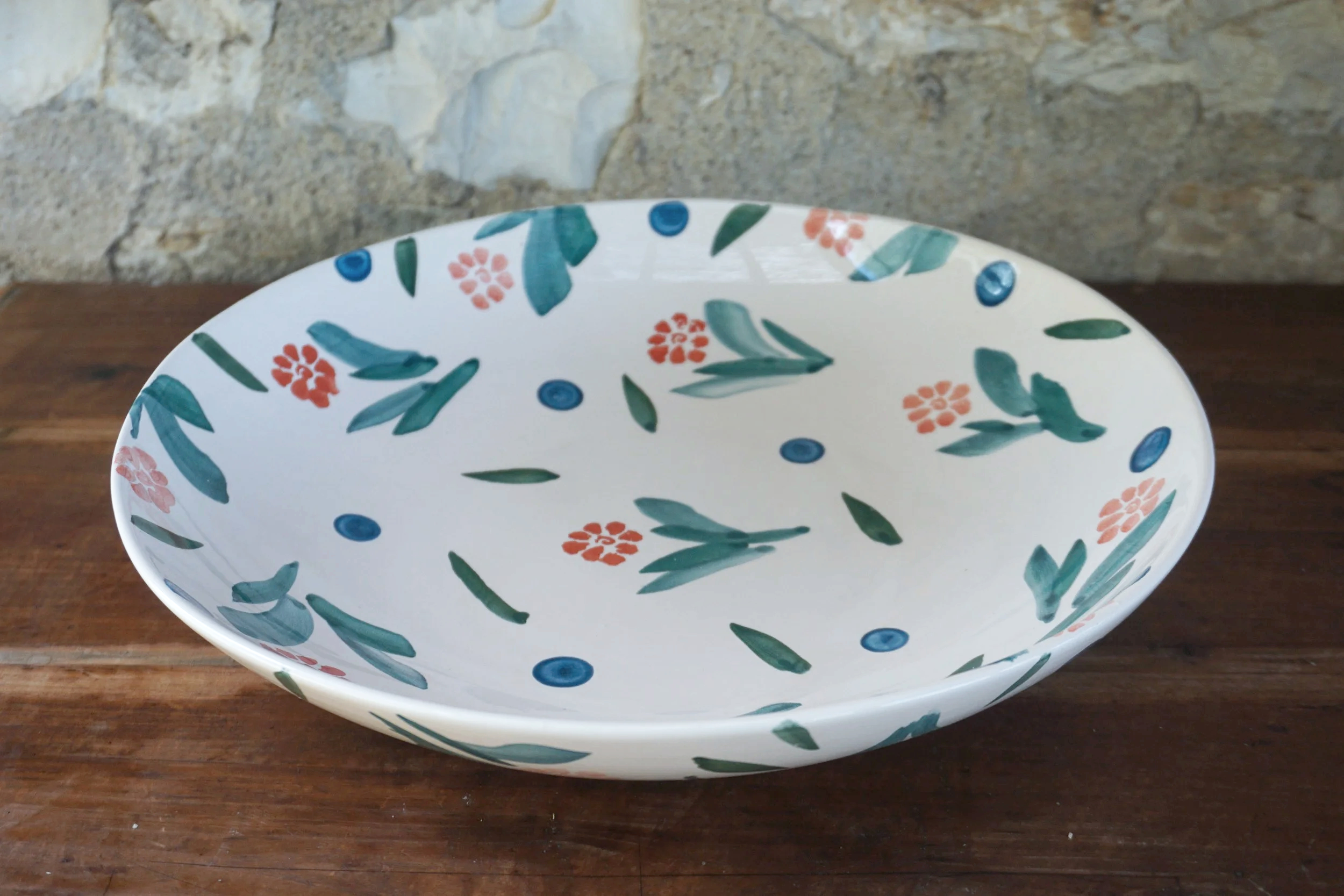 A white ceramic bowl decorated with orange, blue, and green floral patterns, sitting on a wooden surface against a stone wall background.