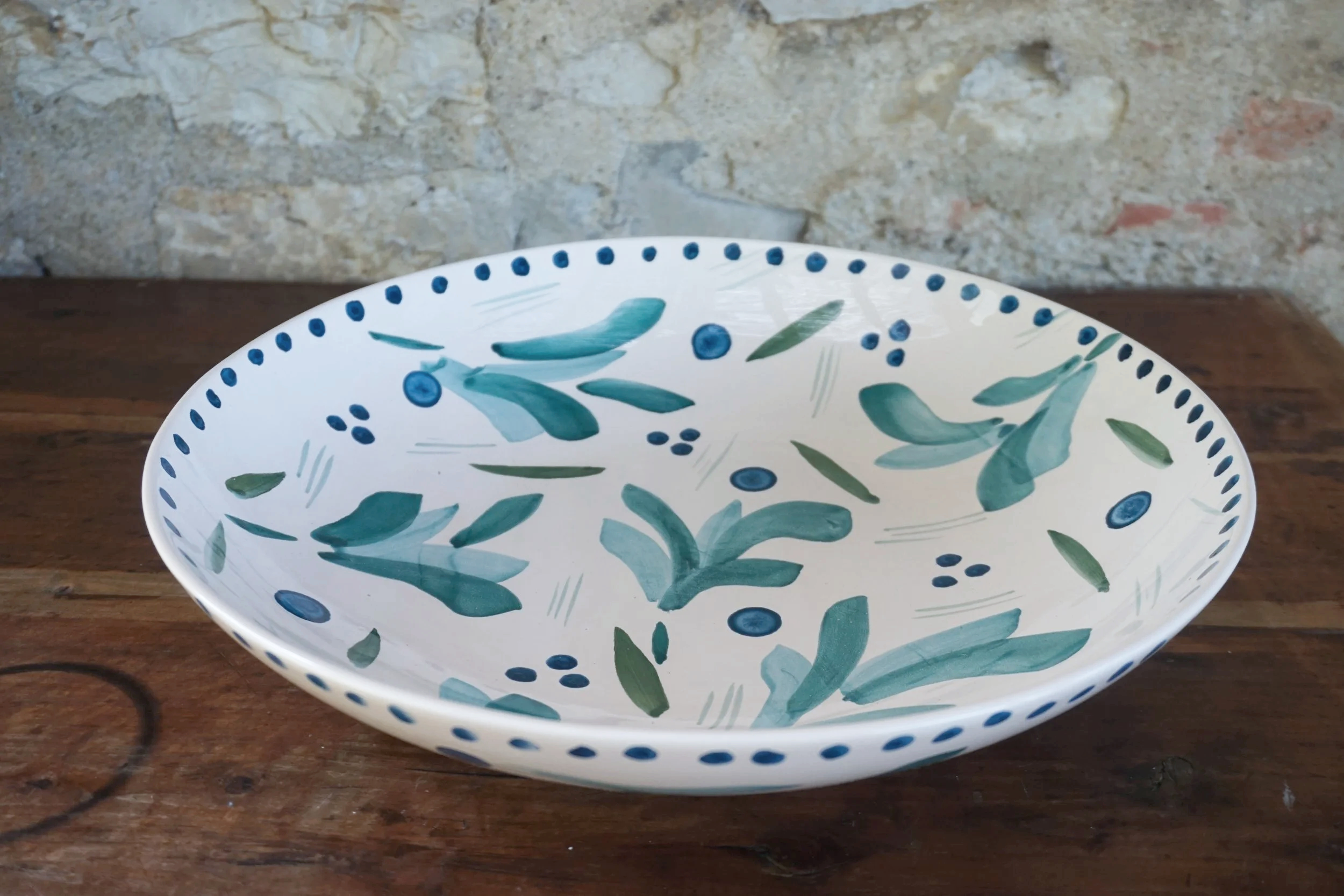 Decorative ceramic bowl with blue and green floral pattern, on a wooden surface, against a stone wall background.
