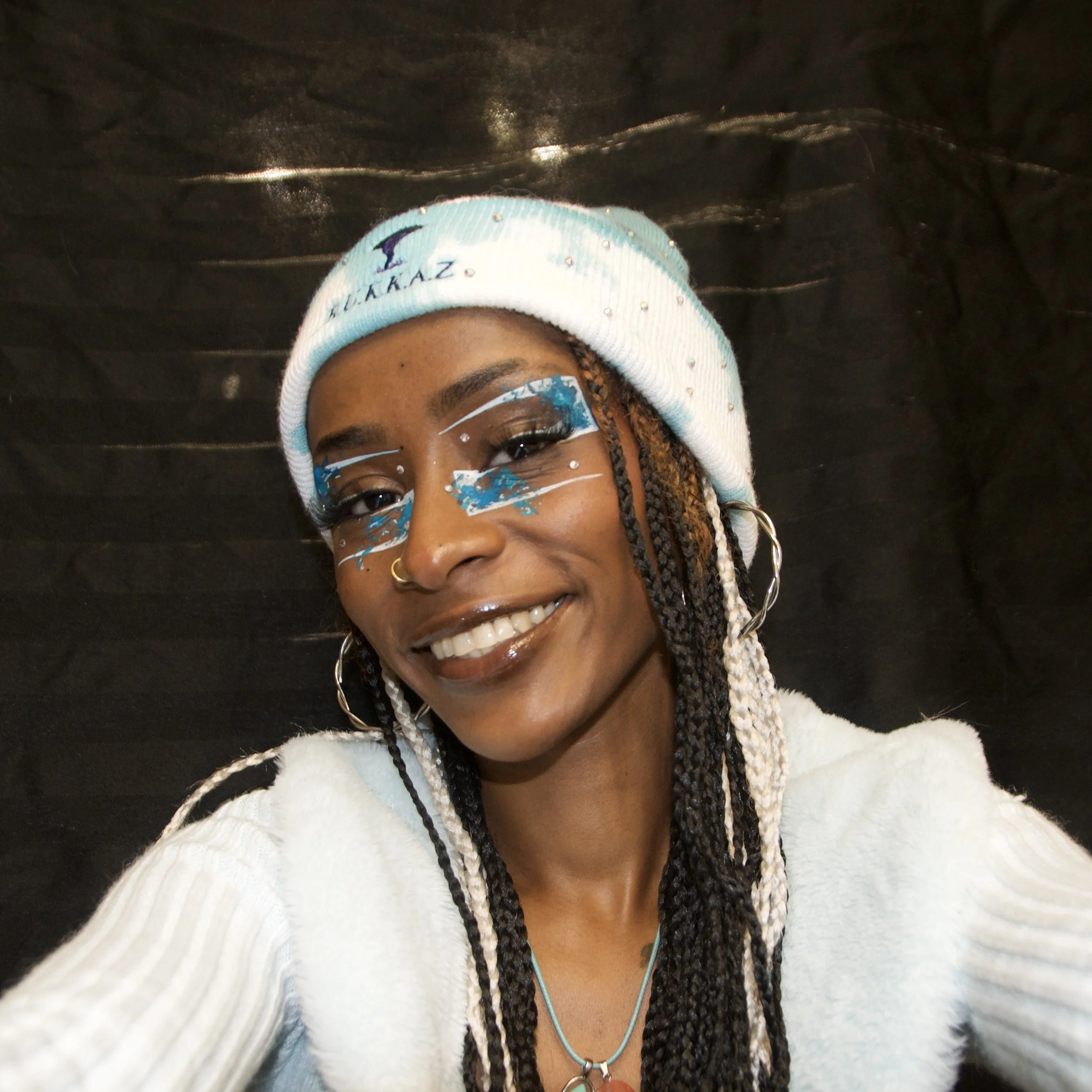 Close-up of a person wearing a white beanie with small embellishments and blue logo, face paint with white and blue designs, and a necklace with a round pendant, against a dark background.