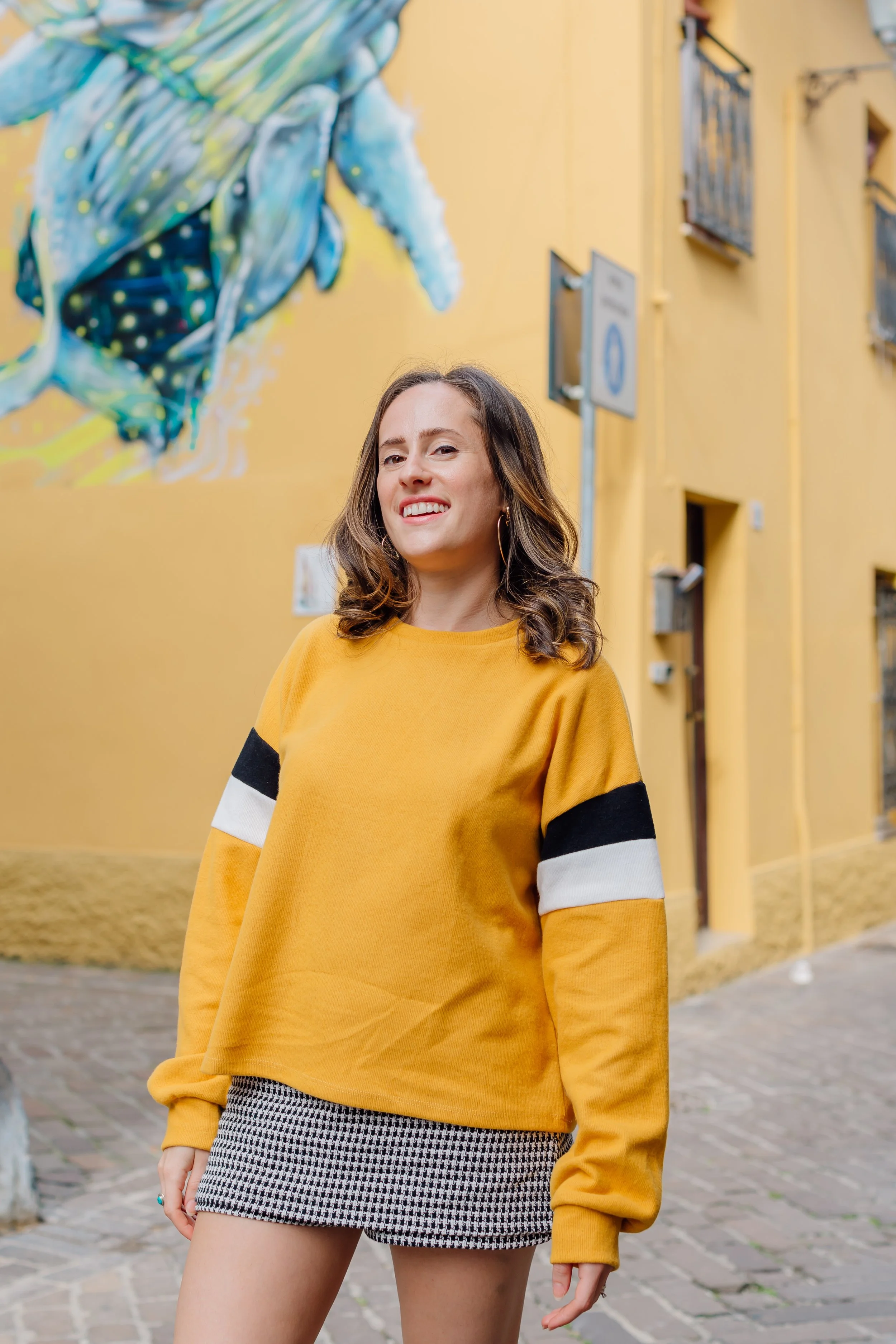 Donna con maglione giallo e gonna a quadri, camminando su una strada pavimentata, con un murale colorato sullo sfondo.
