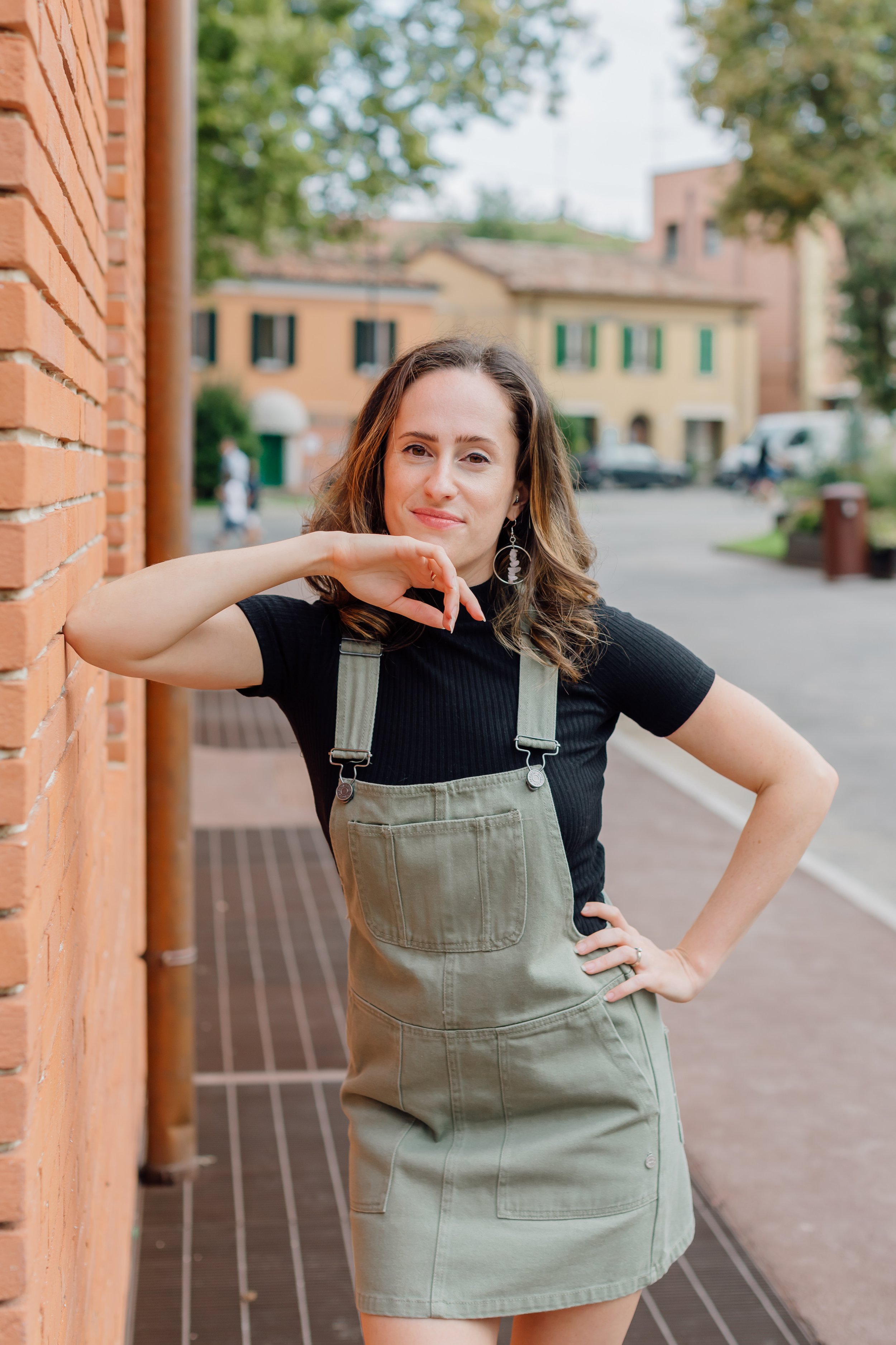 Una donna con capelli castani ondulati, indossa una maglietta nera e un grembiule verde oliva, posa con una mano sul fianco e l'altra vicino al viso, davanti a un muro di mattoni e a un'area urbana con edifici e auto sullo sfondo.