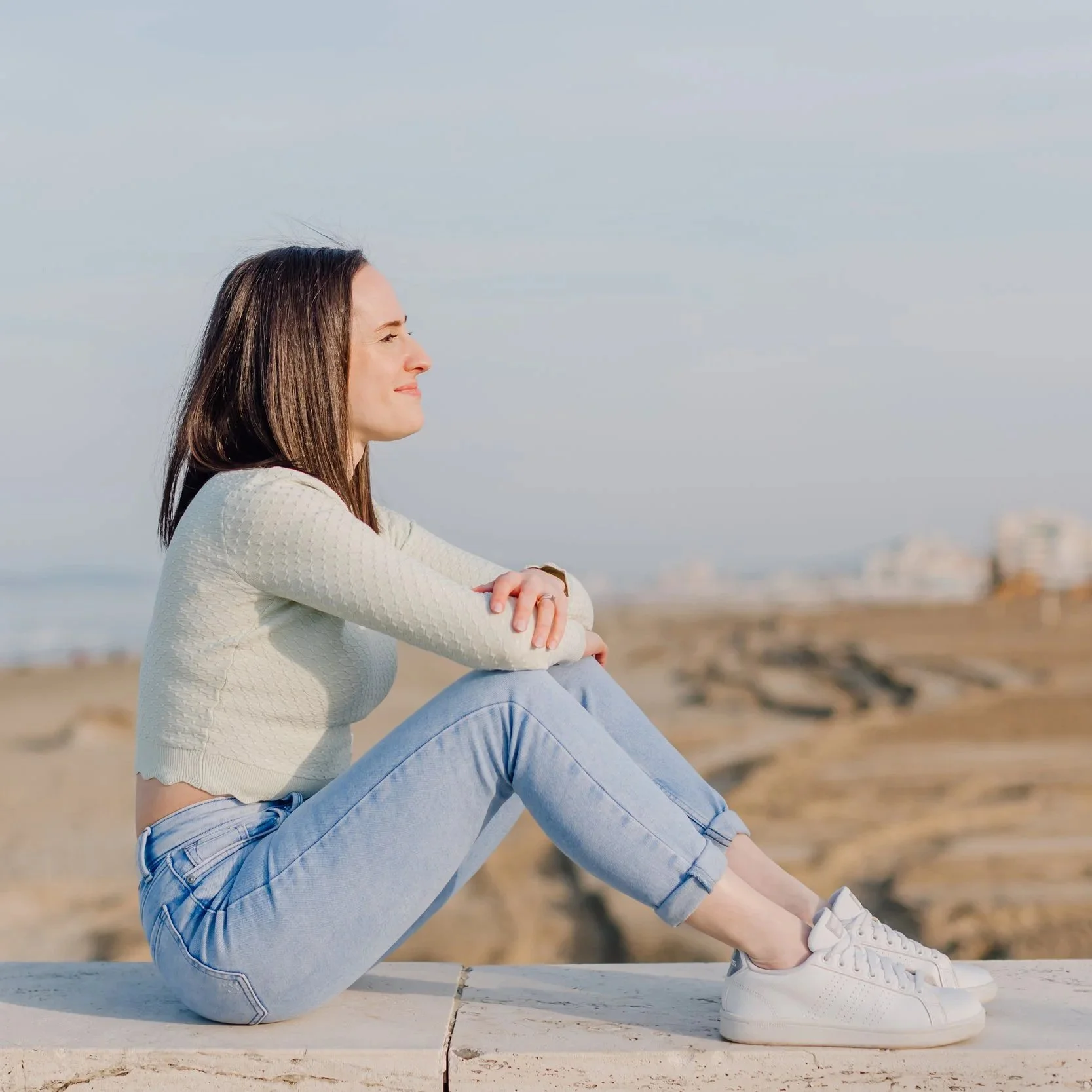 Una donna seduta con le gambe piegate, indossa maglione, pantaloni in jeans e scarpe bianche, guarda verso destra, sullo sfondo un cielo chiaro e una spiaggia.
