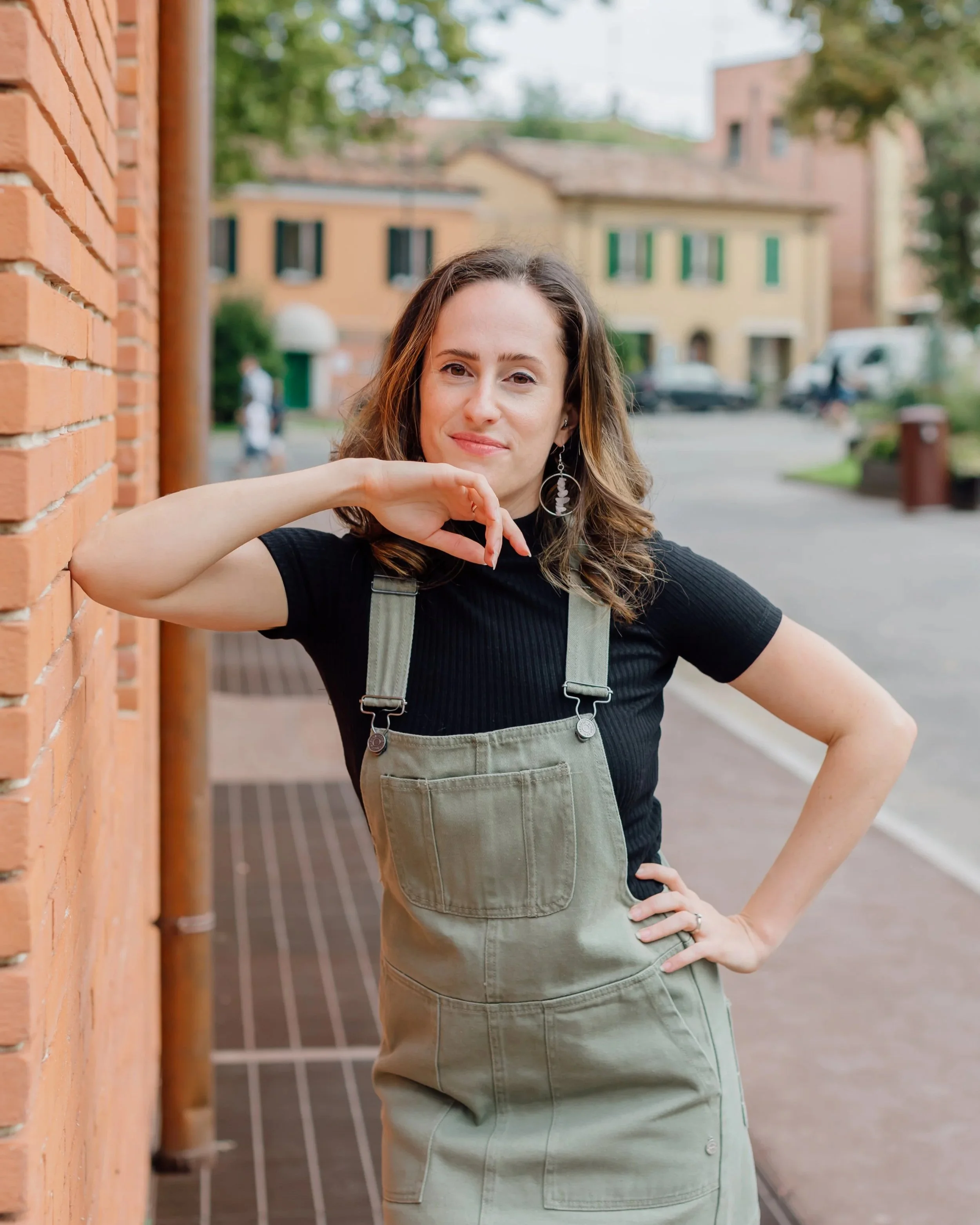 Ragazza con capelli ondulati, orecchini pendenti, vestita con maglietta nera e salopette verde oliva, che sorride e si appoggia alla guancia con una mano davanti a un muro di mattoni rossi, in un contesto urbano con edifici e veicoli sullo sfondo.