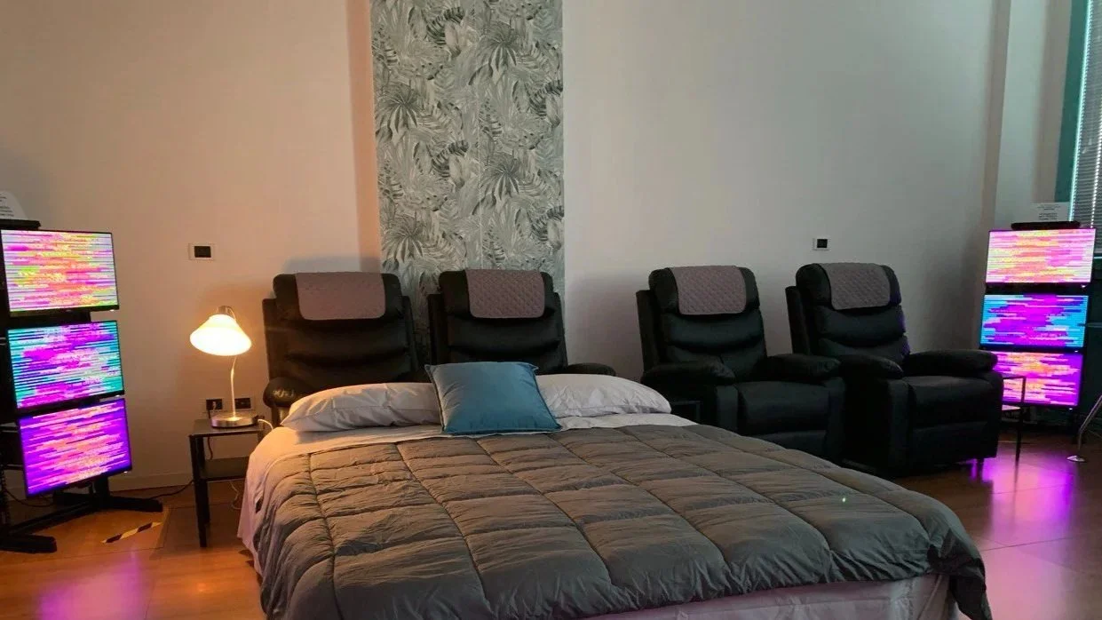 Sala centro olistico con letto matrimoniale, poltrone nere con cuscini, e tecnologia EESystem Light System nei quattro angoli della stanza creando un campo elettromagnetico.