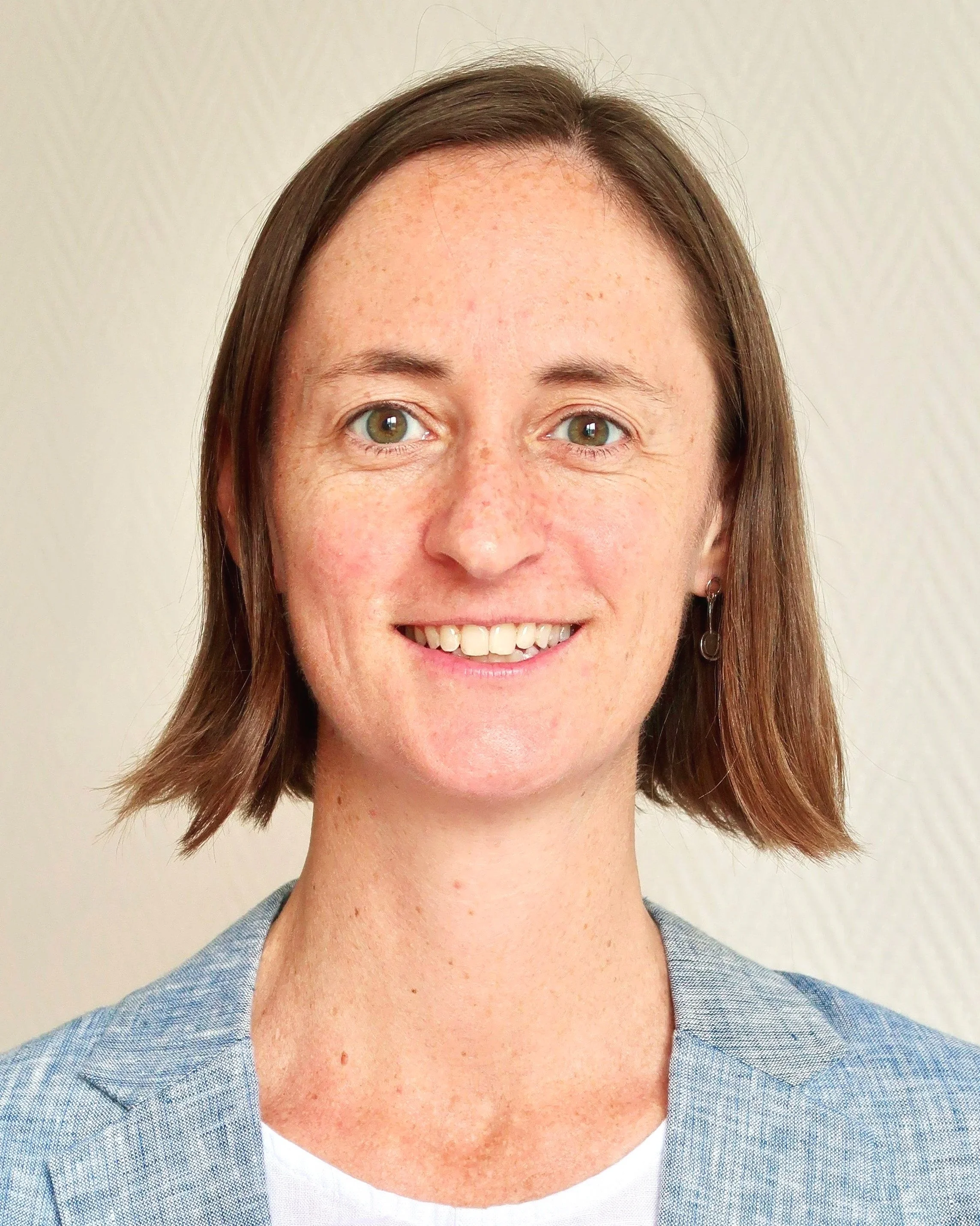 Close-up of Sarah Conway's face showing brown hair, light skin with freckles, hazel eyes, smiling with visible teeth, wearing a light blue blazer and a white top, against a plain light background.