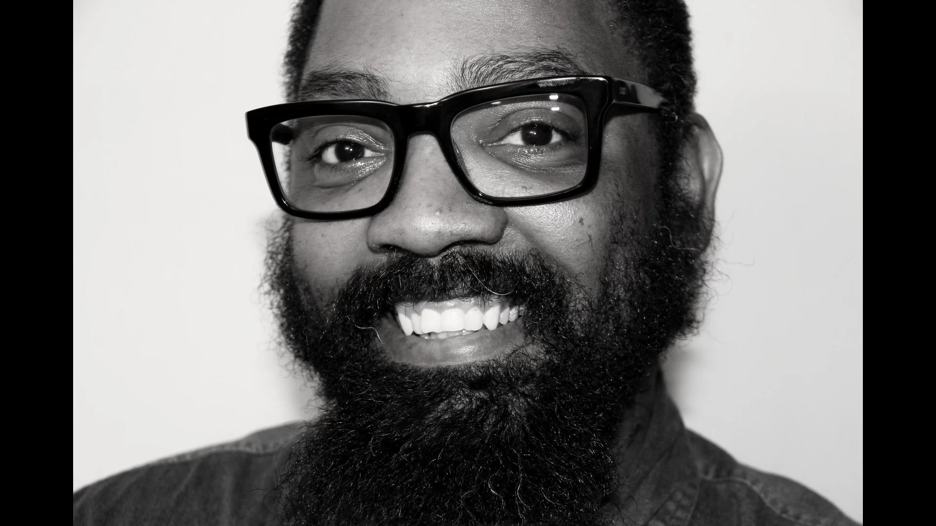 A man with a full beard wearing glasses smiling at the camera in black and white.