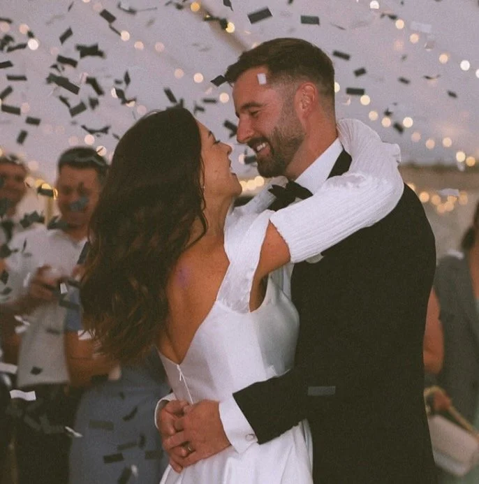 A couple dancing closely at a celebration, smiling at each other with confetti falling around them and string lights in the background.