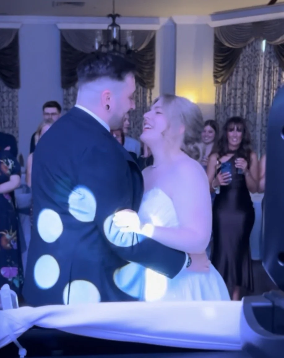A newlywed couple dancing closely at their wedding reception with guests watching and smiling in the background.