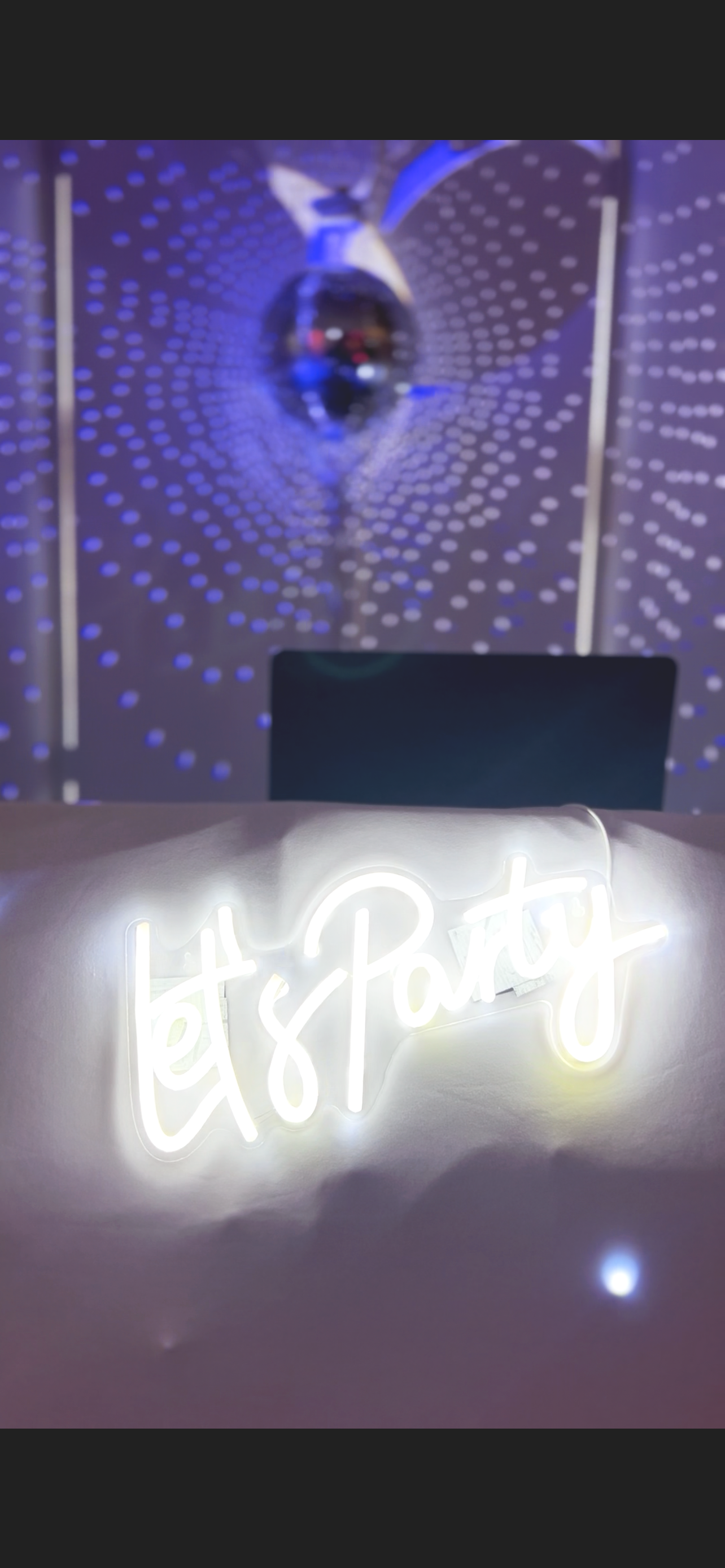 Backlit neon sign spelling 'Let's Party' in cursive, with a blurred background of dotted blue lights and a reflective metallic object.