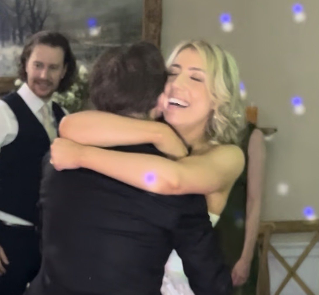 People hugging at a celebration or special event, with one woman smiling happily and a man embracing her, others in the background smiling.