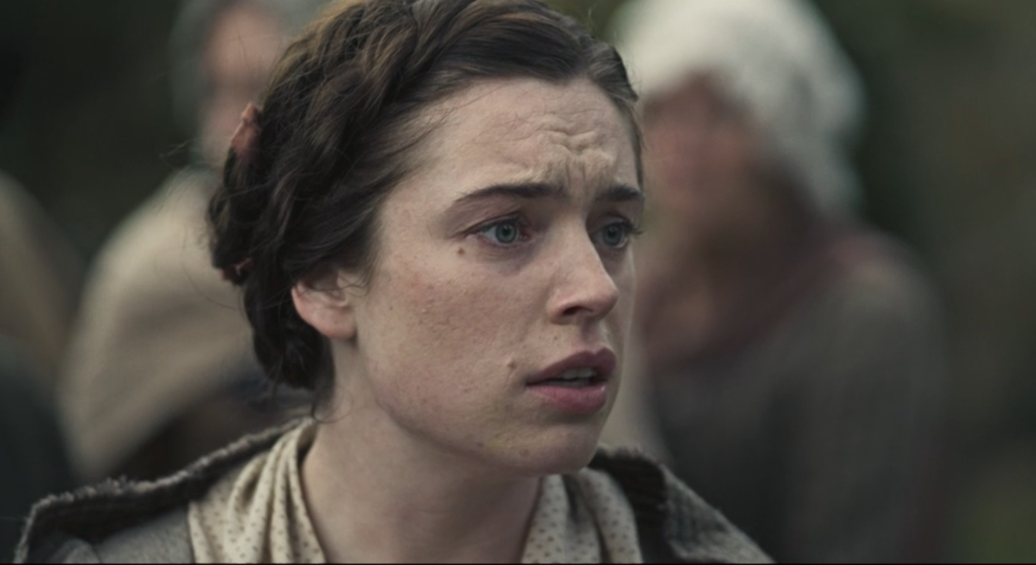 Close-up of a woman with short braided dark hair, light complexion, and blue eyes, with a concerned or distressed expression, in a scene with a blurred background of people.