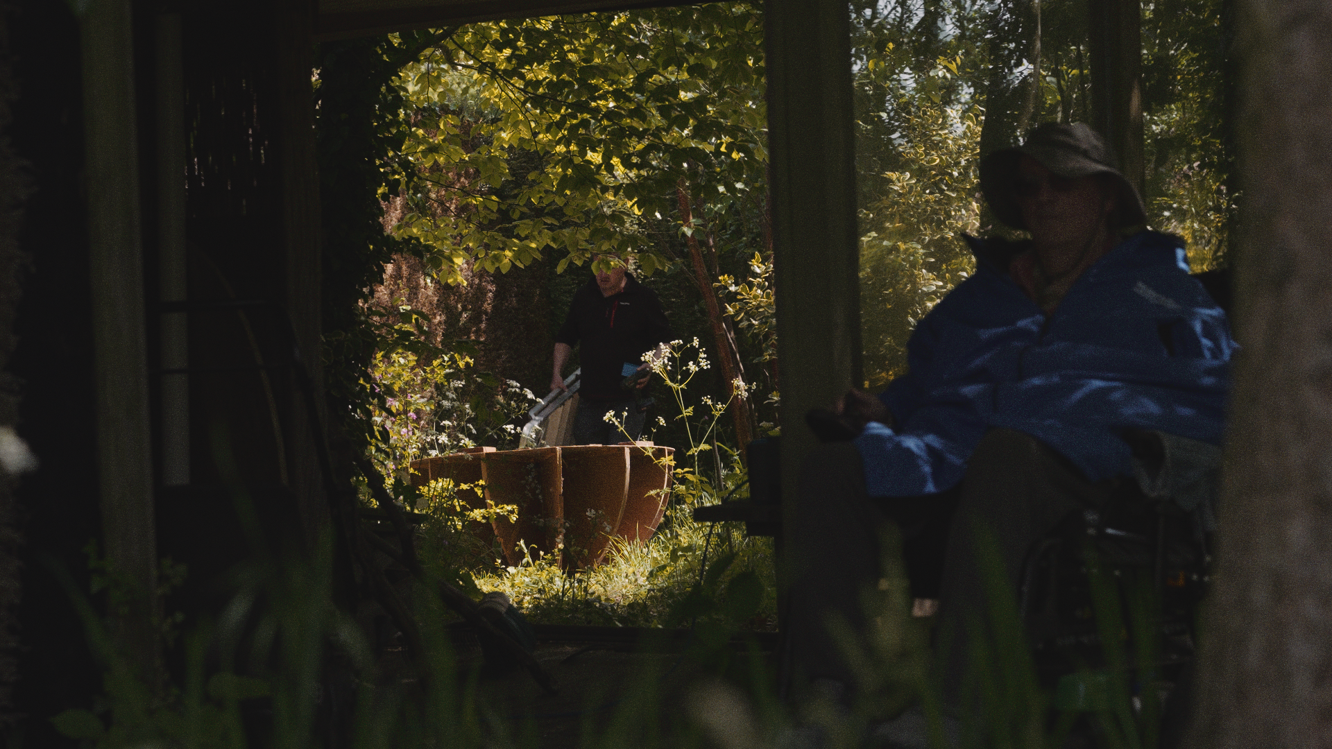A person sitting in a wheelchair under a shaded area, with two other people in the background in a lush outdoor garden, near a pond or water feature.