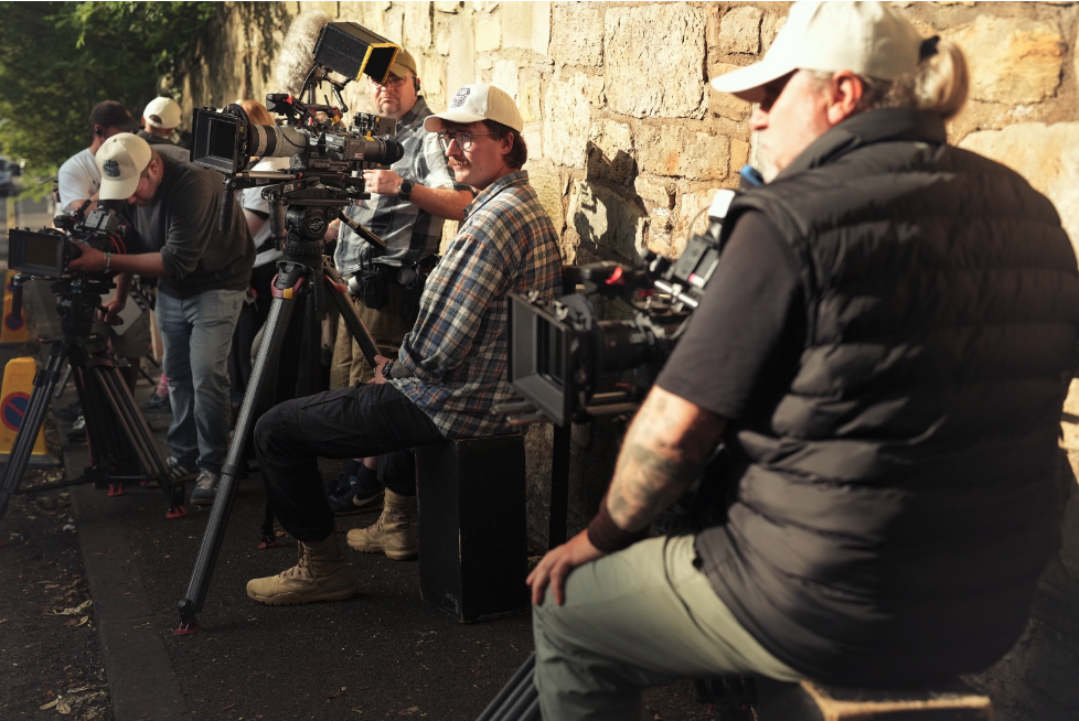 Group of filmmakers or videographers with cameras and equipment filming outdoors against a stone wall.