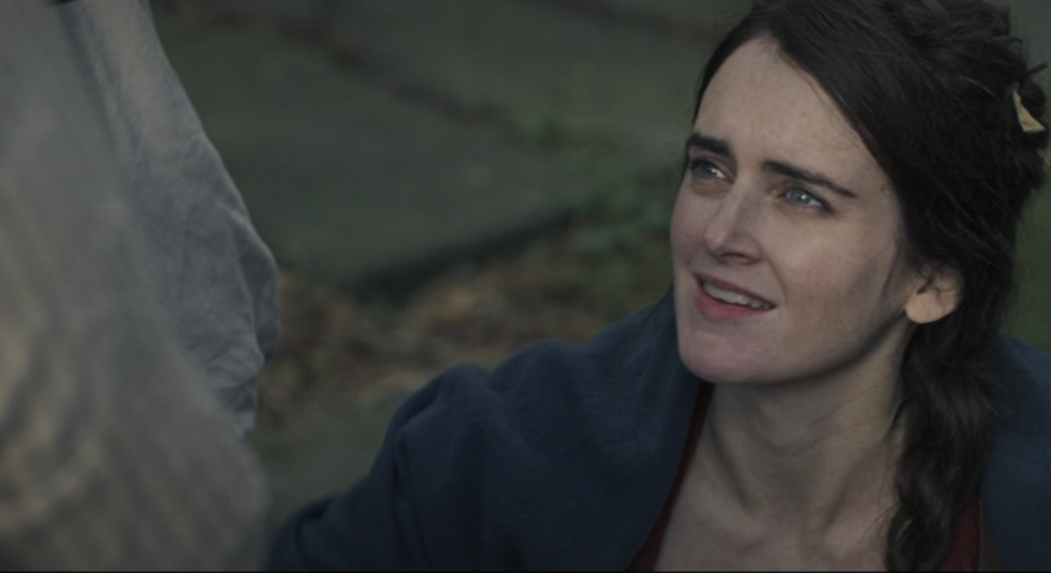 A woman with dark hair and light skin looks up with a smile, outdoors with greenery behind her.