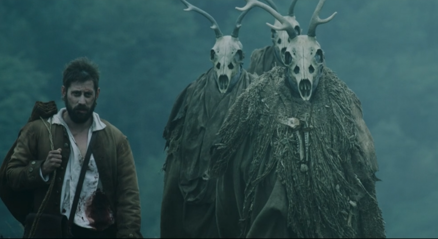 A man walking in a forest with two large mythical creatures wearing skull masks and antlers.
