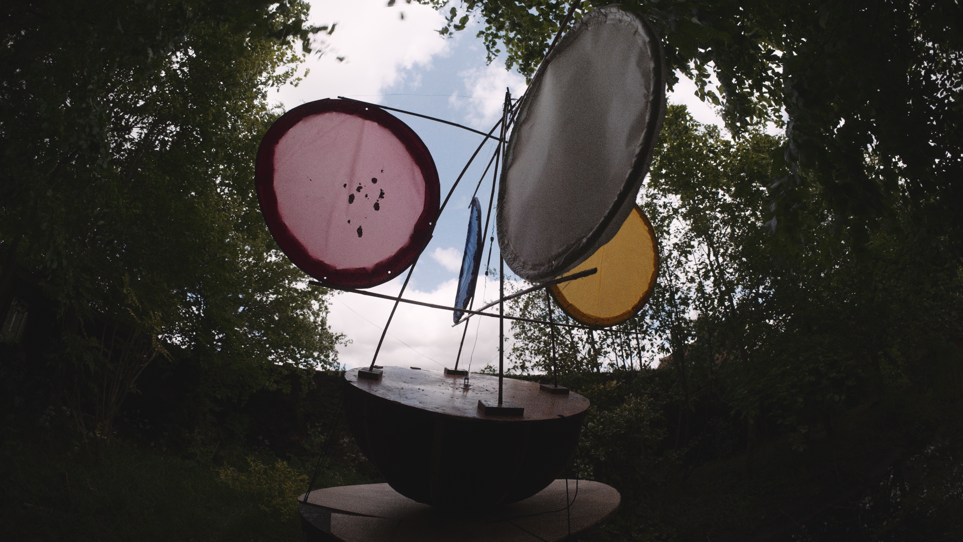 Outdoor sculpture of a boat with three large circular objects resembling drum surfaces, surrounded by trees and under a cloudy sky.