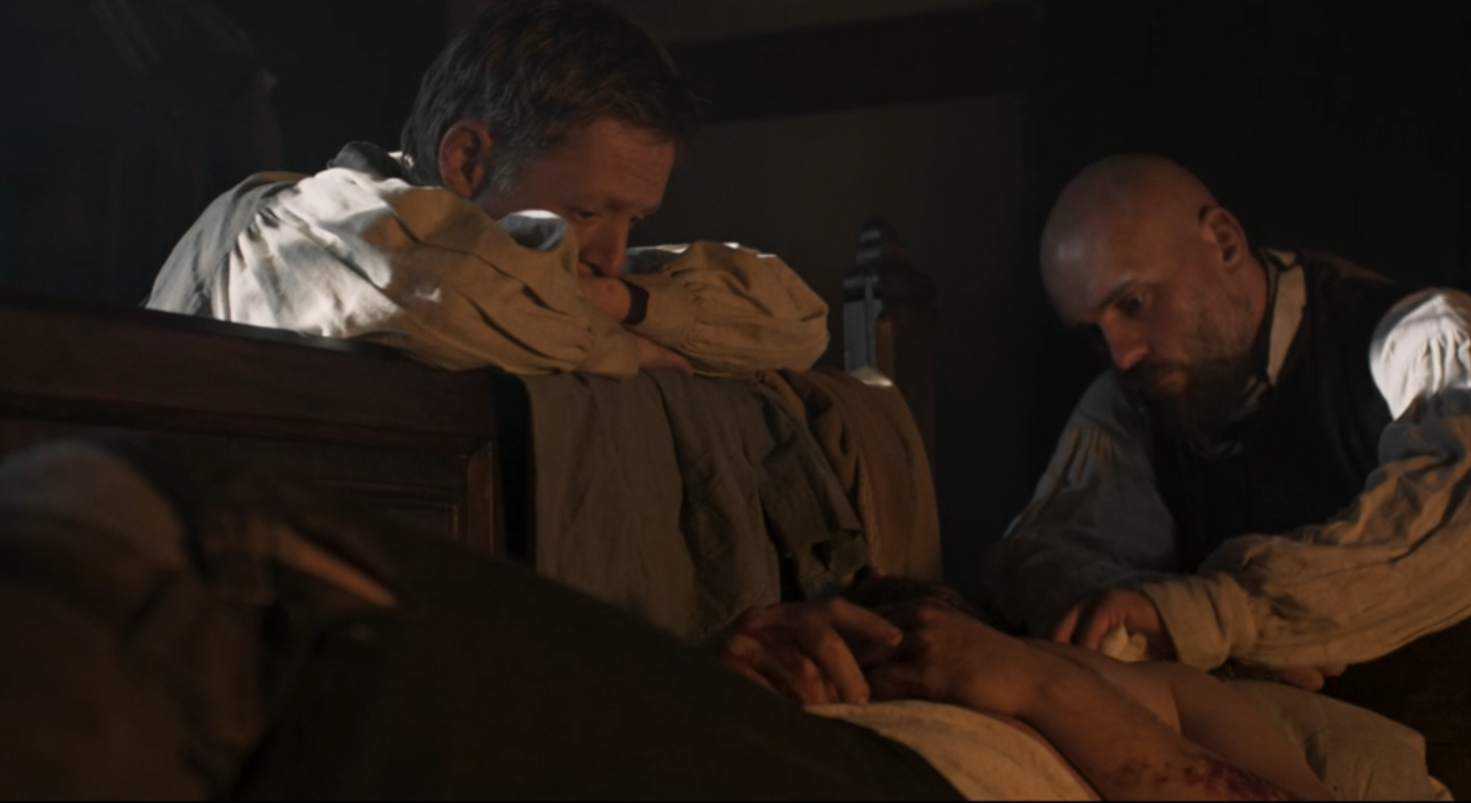 Two men in period clothing kneel beside a bed, holding a person's hand in a dimly lit room.