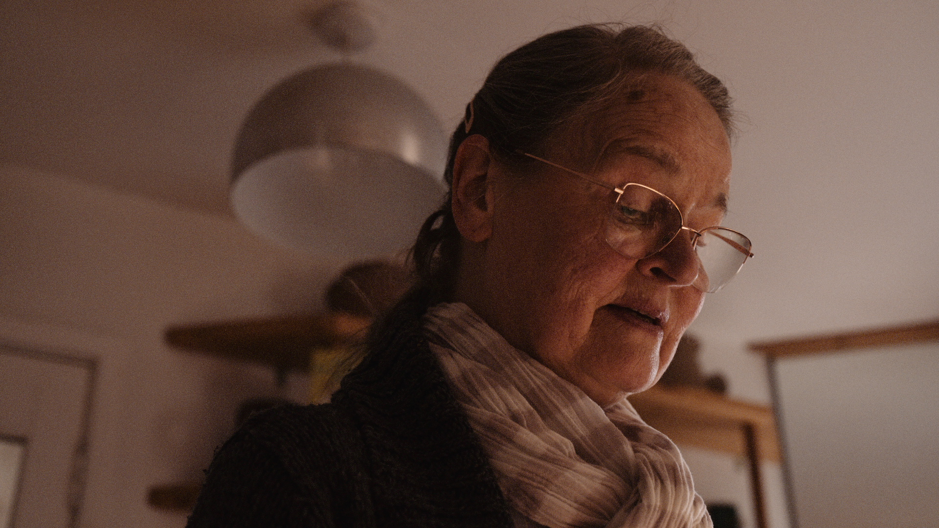 An elderly woman with gray hair and glasses, wearing a striped shirt and scarf, looking downward in a warmly lit room.